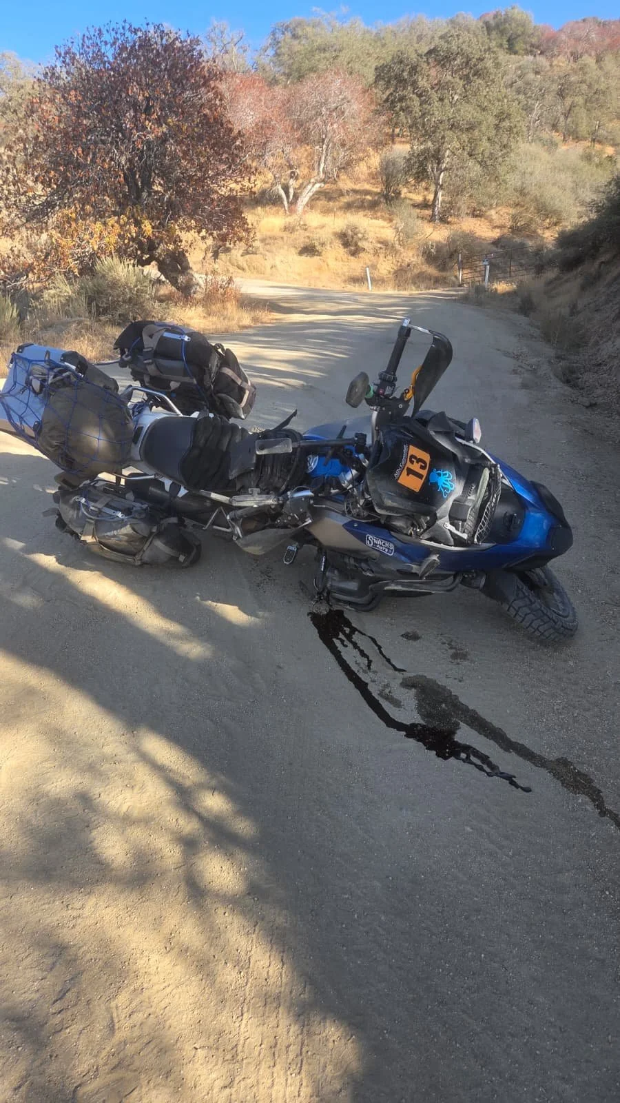 A blue BMW numbered 13 lying on its side on a dirt mountain road with a dark streak of oil running downhill from it