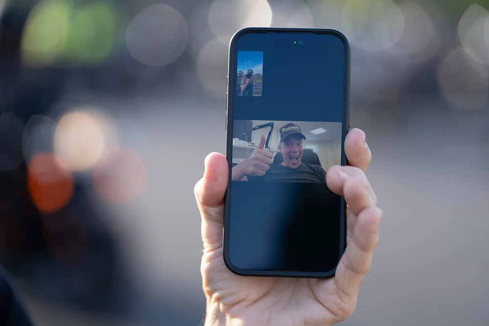 A hand holding up a phone showing Robert Baldinger giving a thumbs up on a video call