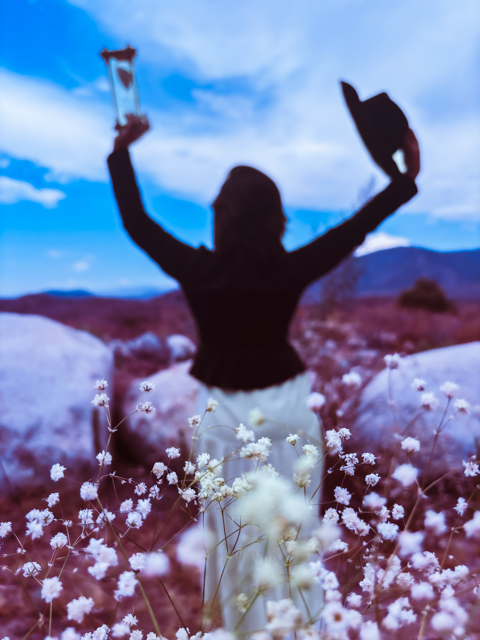 Silhouette of a woman standing in a field of white flowers with her arms raised, holding a hat and a small object, against a blue sky with clouds and mountains in the background.