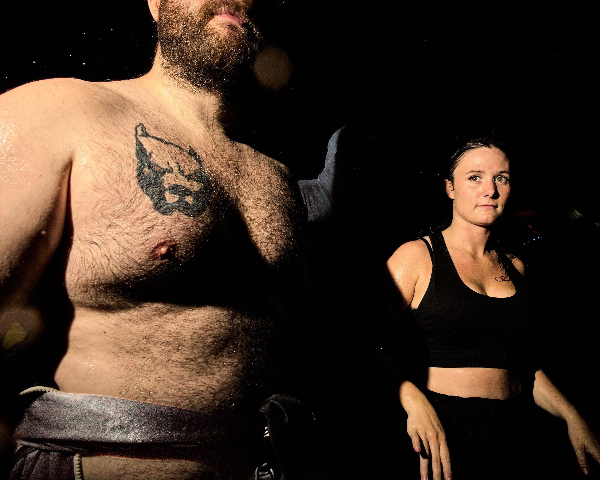 A shirtless man with a beard and chest hair, and a tattoo of a bear on his chest, standing next to a woman with dark hair wearing a black sports bra. Both are against a dark background.