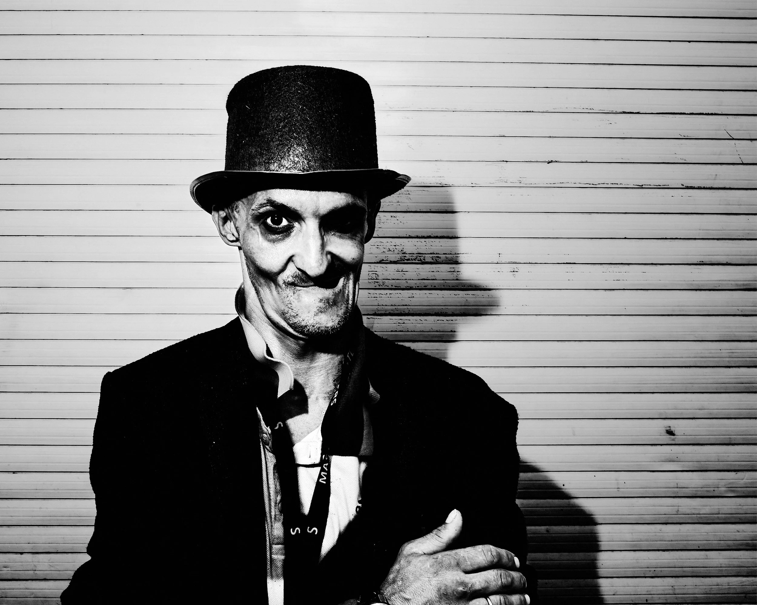 Black and white photo of a man with dark hair, wearing a black top hat and a dark jacket, smiling, against a background of horizontal wooden planks.