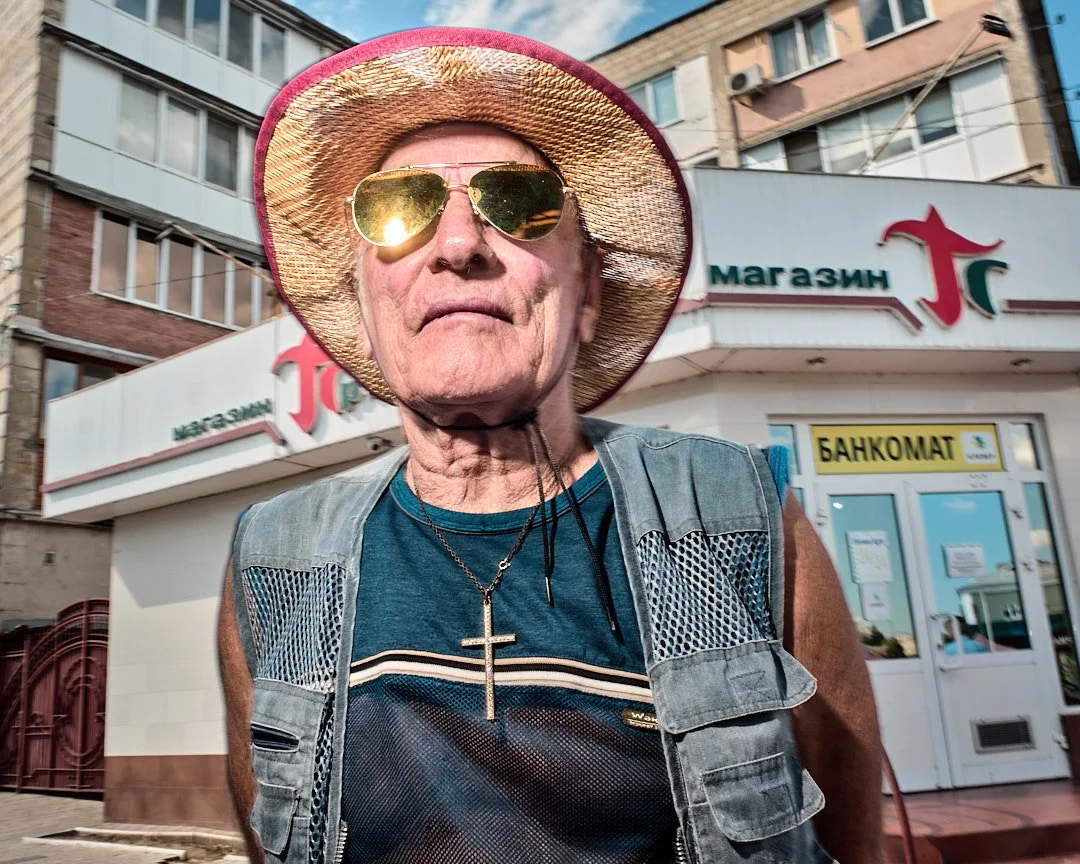 An elderly man wearing sunglasses, a wide-brimmed straw hat, a sleeveless vest, and a necklace with a cross, standing outside a store with Cyrillic signage.
