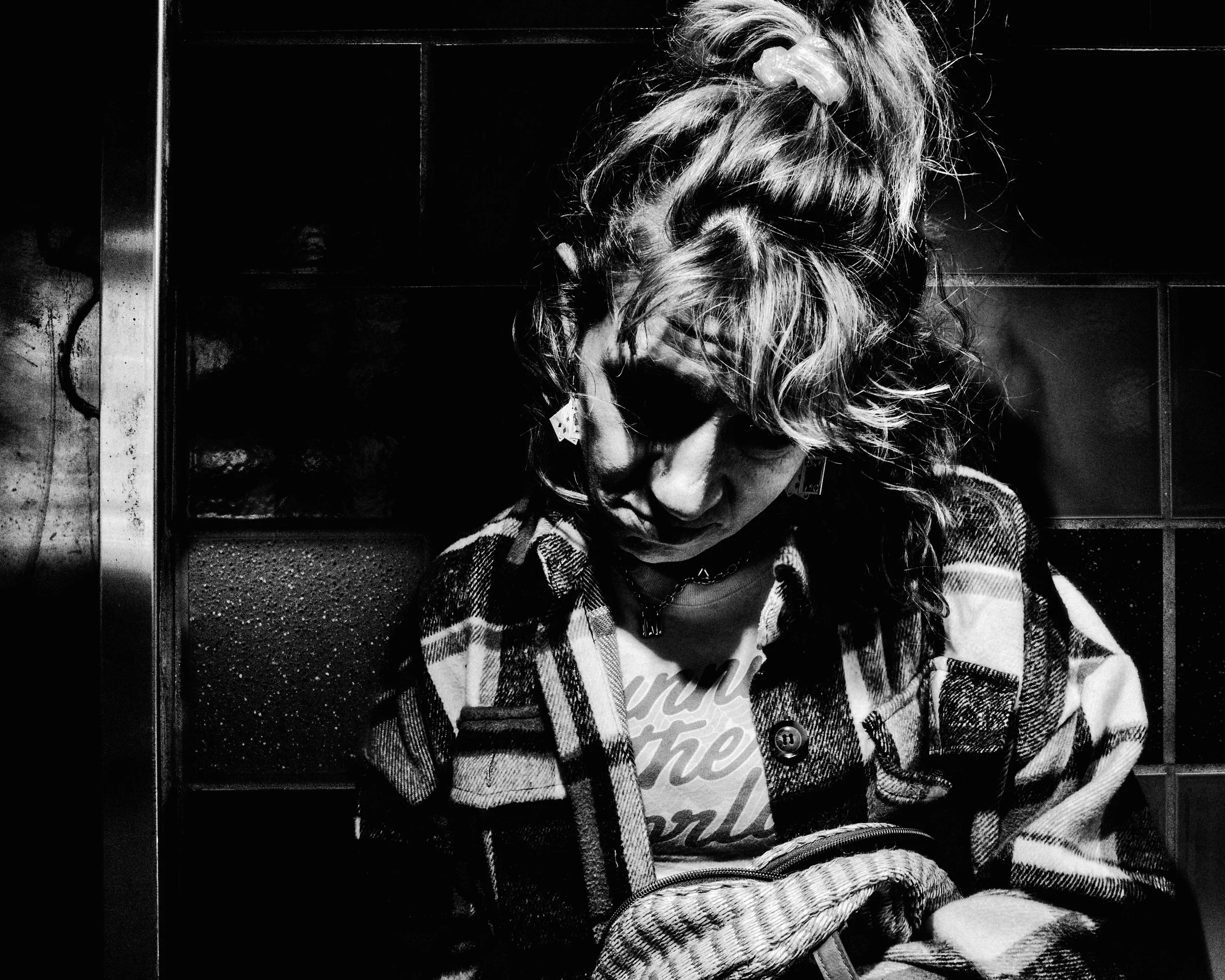 A young woman with curly hair tied in a high ponytail with a scrunchie, wearing a plaid shirt, looking down, standing in front of dark lockers.