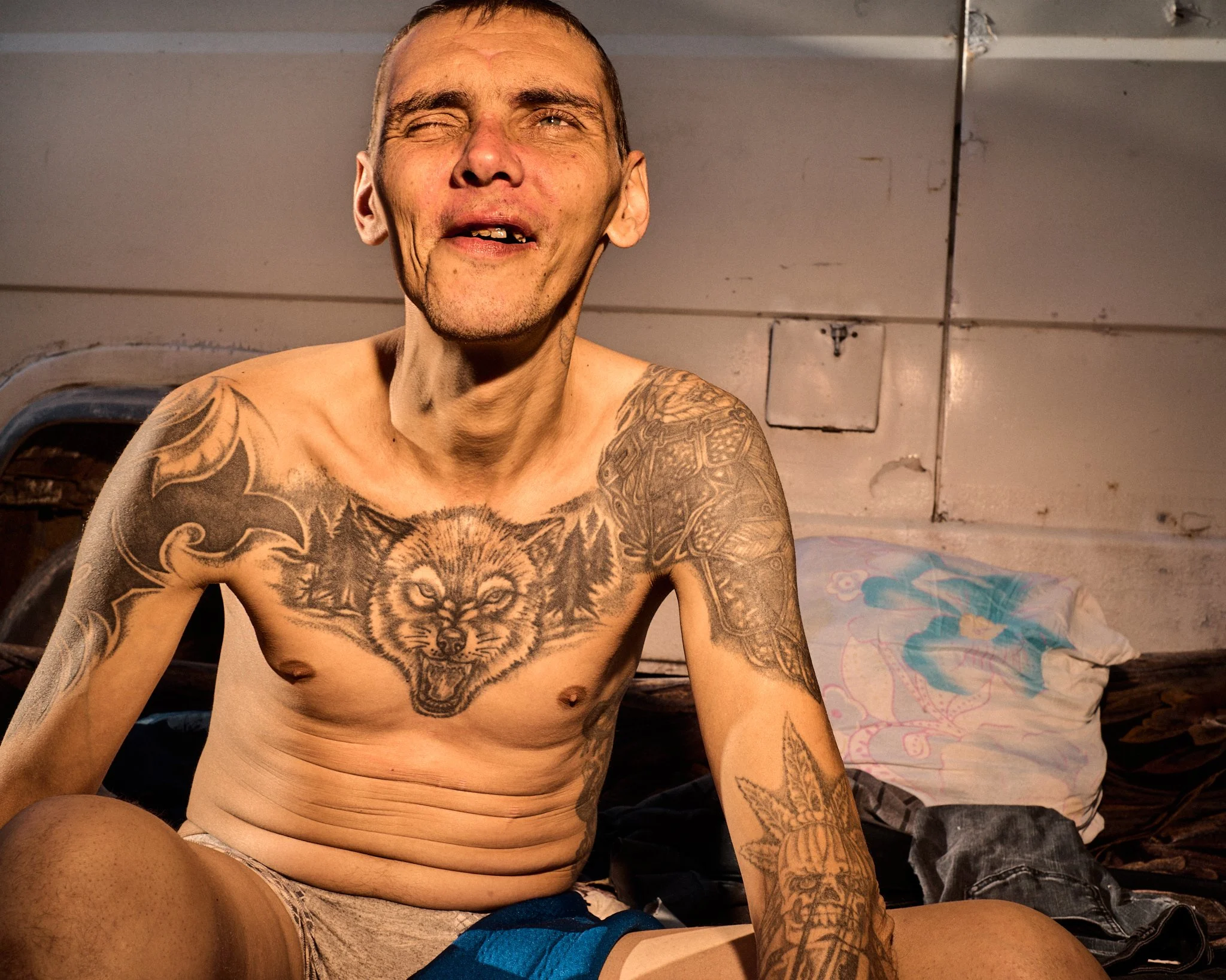 A shirtless man with tattoos sitting on a bed in a rustic setting with a pillow and clothes in the background.