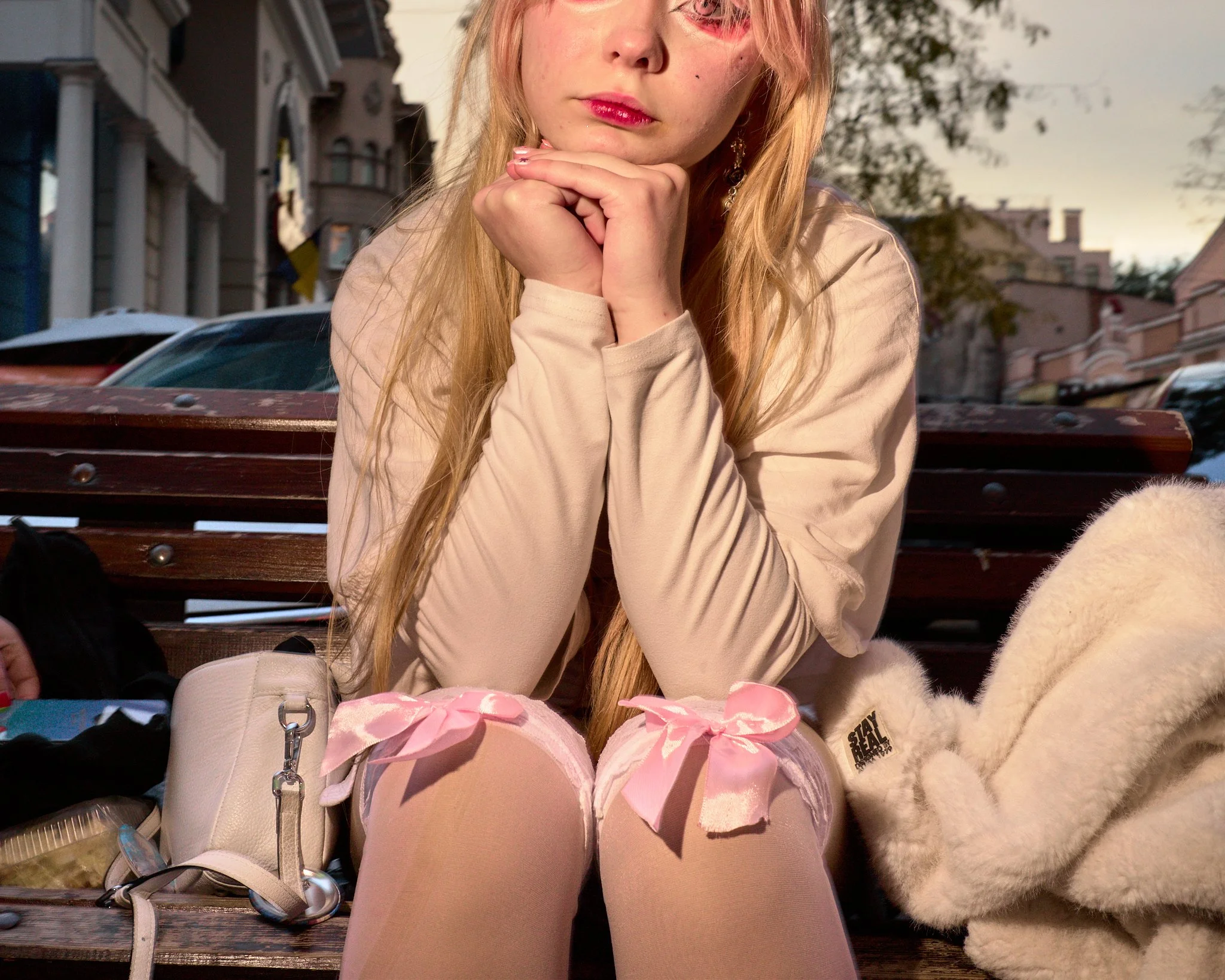 A young woman sitting on a park bench with her chin resting on her hands, wearing a beige jacket and pink knee-high socks with bows. There is a white fuzzy coat and white purse nearby, and a dog lying next to her. Background includes residential buildings and trees at sunset.
