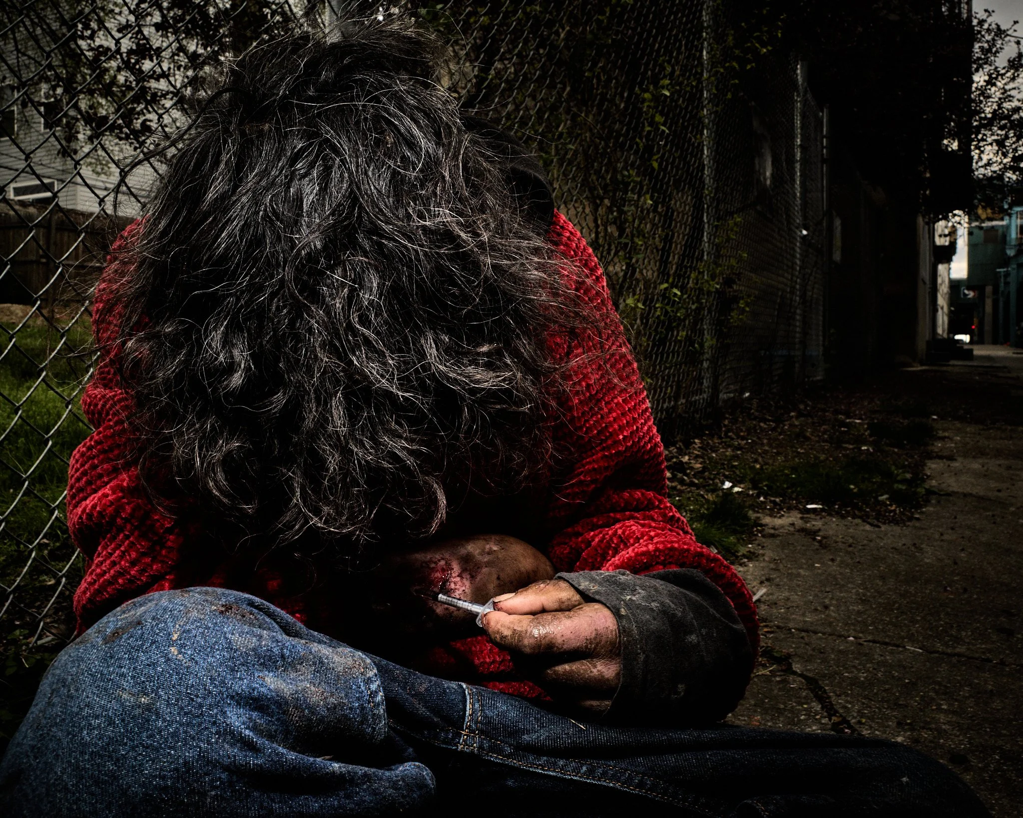 A homeless person with long, curly gray hair sitting on the ground, holding a syringe, near a chain-link fence on a dark street.