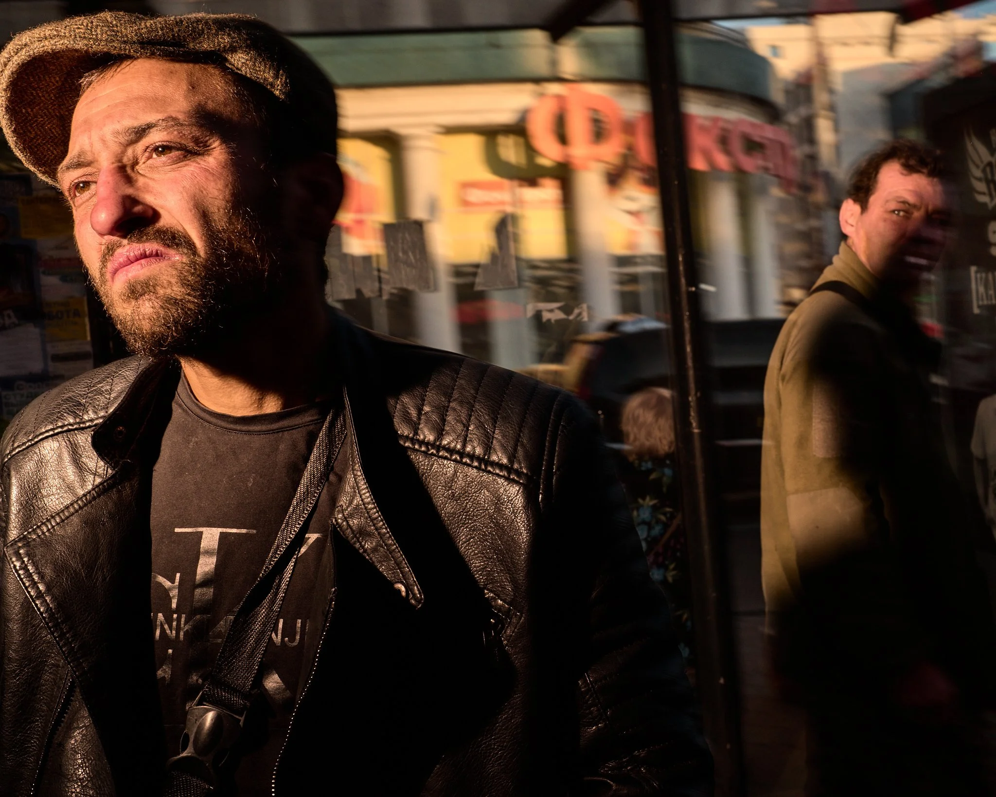 A bearded man wearing a leather jacket and a hat, standing outside and looking to the side. Reflection of another man in a jacket is visible on the glass window beside him. The background includes a storefront with signs and a street scene.