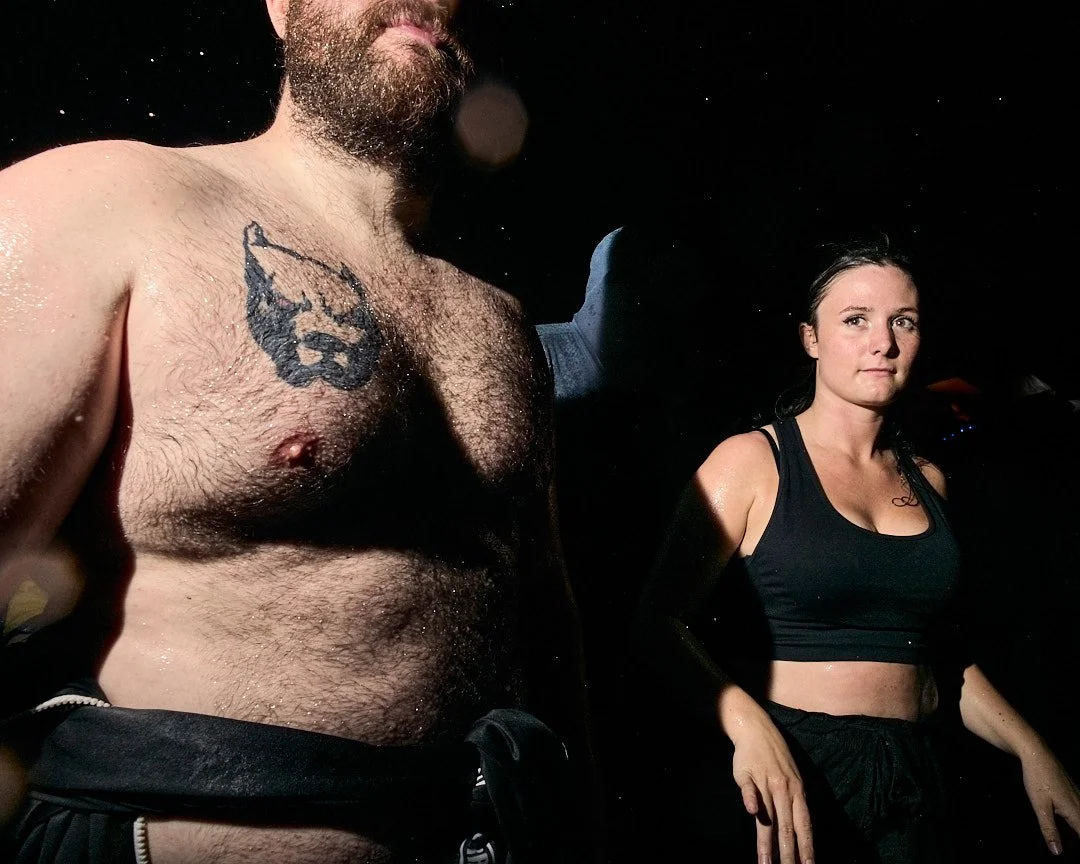 A shirtless man with a beard and a tattoo on his chest stands next to a woman in a black crop top. The background is dark with small points of light, possibly stars or lights.