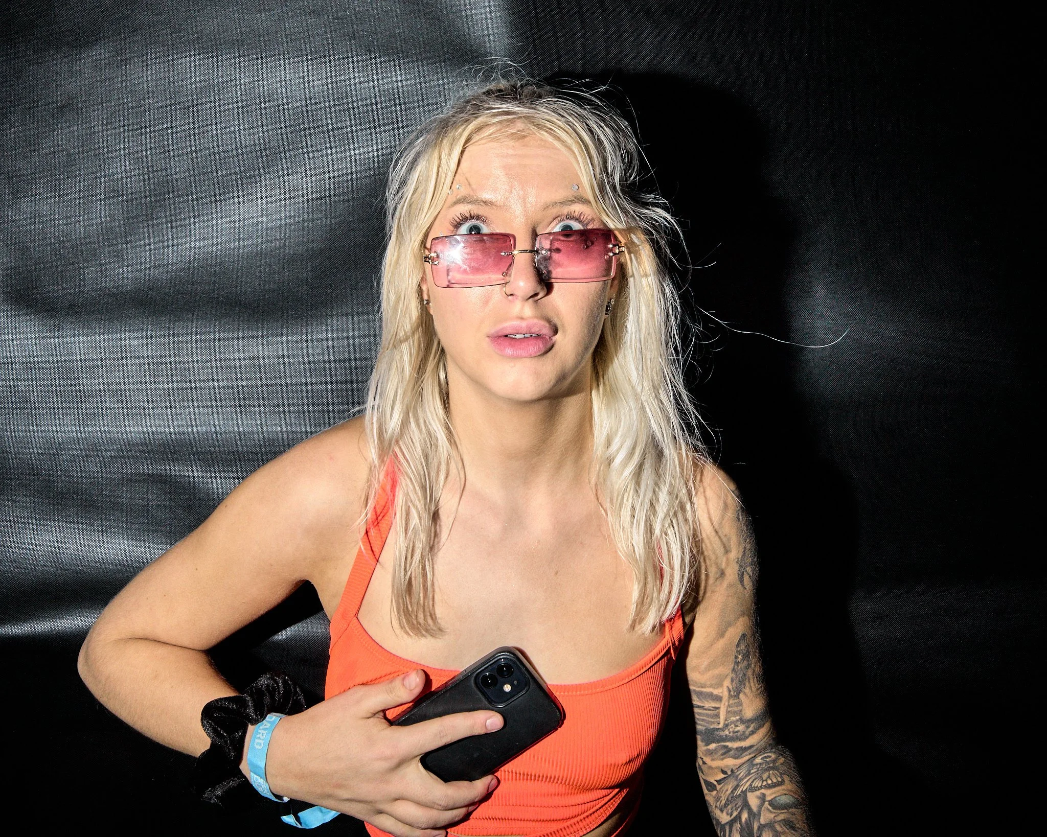 A woman with blonde hair wearing pink-tinted sunglasses, an orange tank top, and tattoos on her left arm, looking surprised while holding a smartphone.