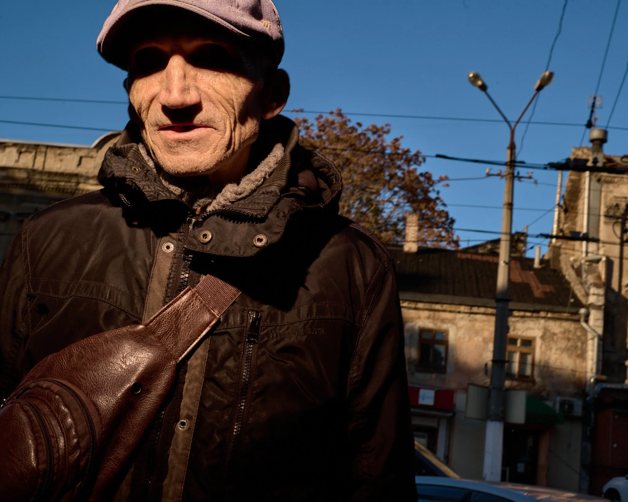 An elderly man with a weathered face, wearing a cap, sunglasses, and a dark jacket, standing outdoors during daytime with utility poles and old buildings in the background.