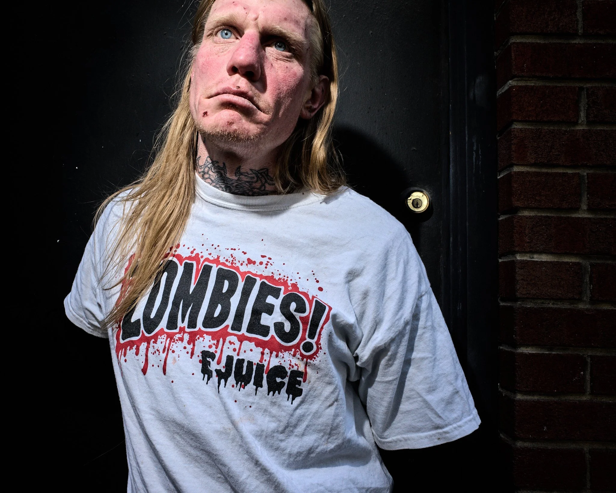 A man with long blond hair, tattoos, and a white T-shirt that says "Zombies! Juice" in blood-spattered font, standing outdoors against a dark background and brick wall.