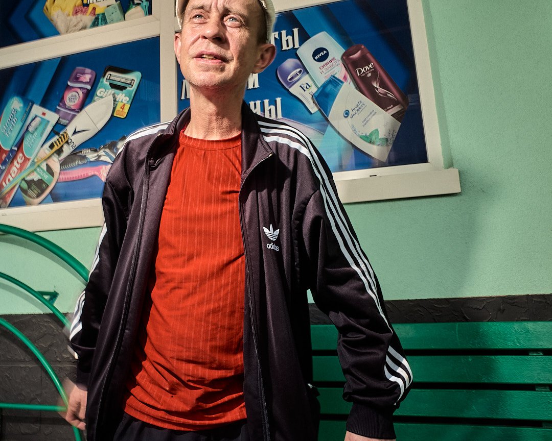 A middle-aged man with light skin and short gray hair, wearing a black Adidas jacket with white stripes and a red shirt underneath, standing indoors against a green wall and a green bench. Behind him, there is a display window with various toiletries including razors, toothbrushes, and bottles.
