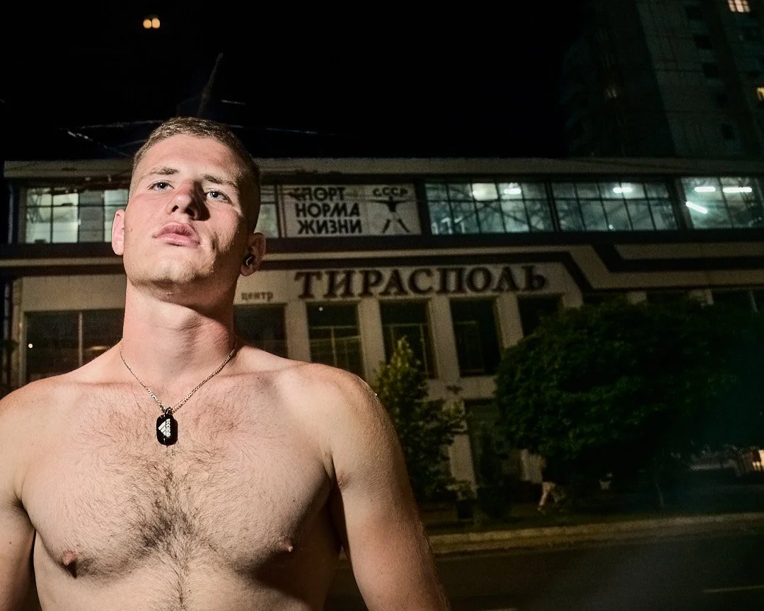 A shirtless young man with short hair and a necklace stands outside at night, in front of a building with large glass windows and a sign in Russian.