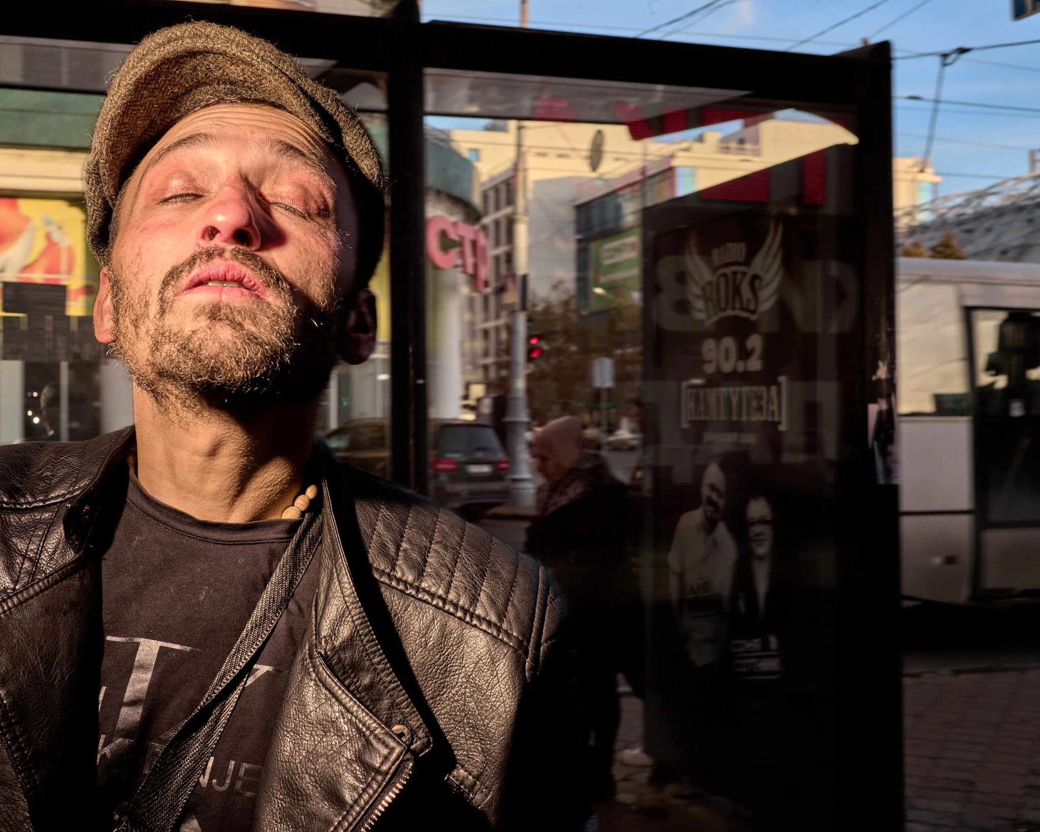 A man with a beard, wearing a leather jacket and a beaded necklace, stands outdoors with his eyes closed and face tilted upward in sunlight. Behind him is a bus stop with reflections of a street scene and two people walking, with a cityscape and buildings visible in the background.