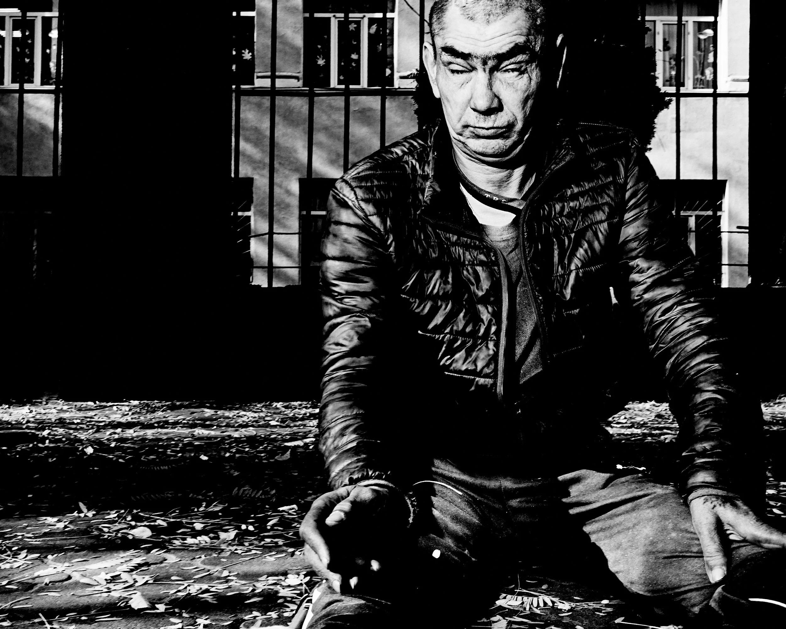 A black and white photo of a man sitting outdoors, wearing a puffy jacket. He appears to be looking downward with a serious expression, with a fence and building in the background.