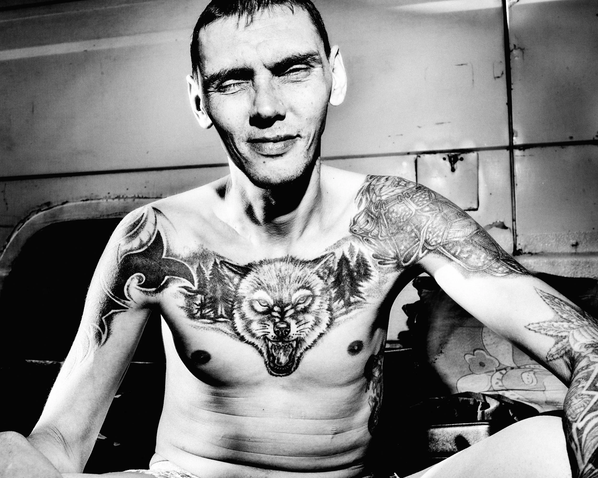 Black and white photo of a shirtless man with tattoos on his chest and right arm, smiling with eyes closed, against a plain background.
