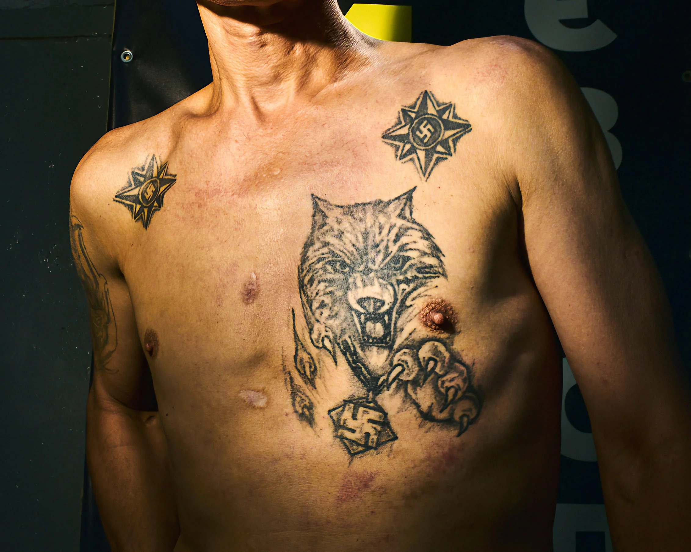 A person's upper torso with several tattoos, including two stars with swastikas on the shoulders and a detailed wolf face on the chest.