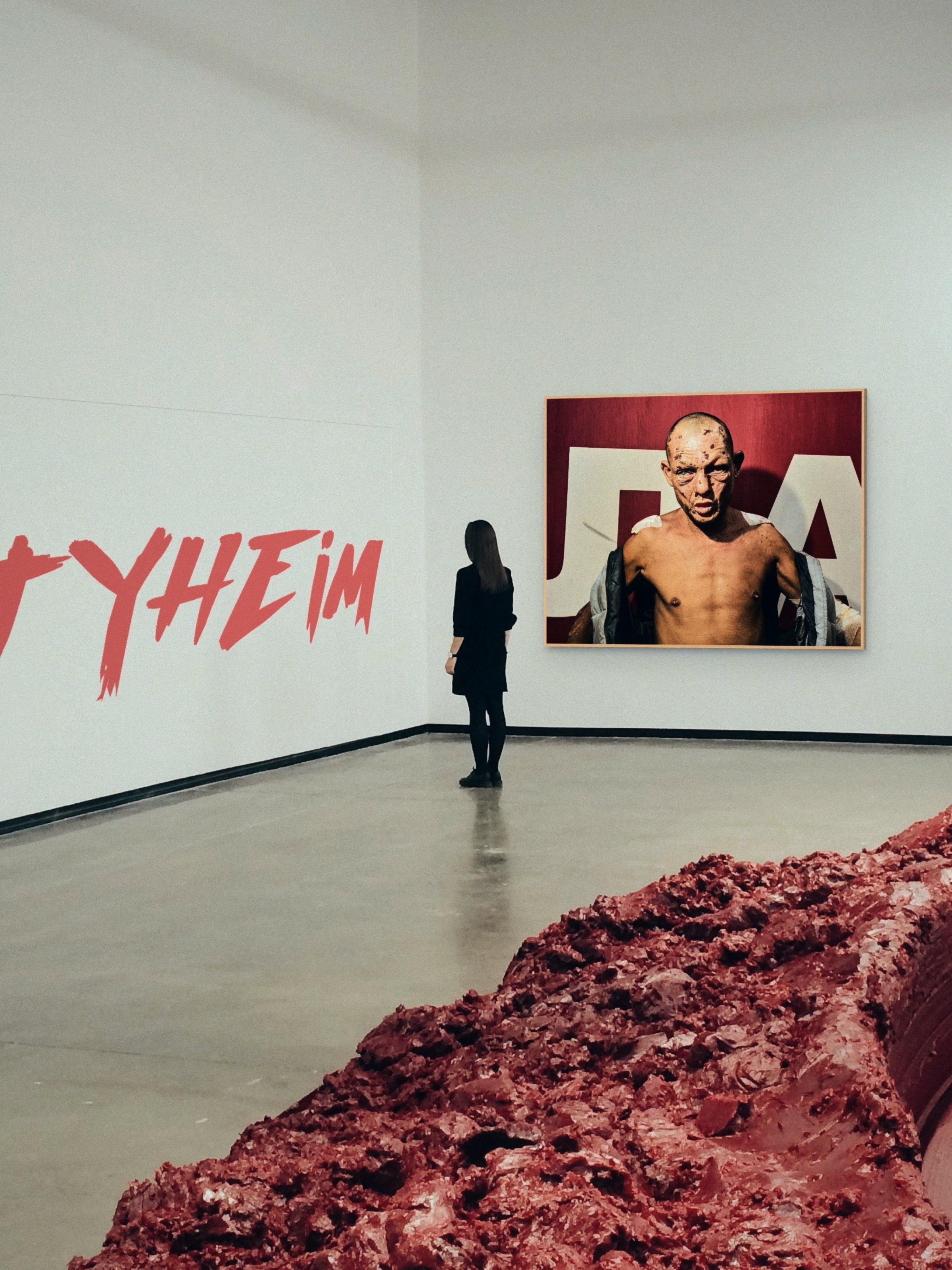 An art exhibition with a large portrait of a topless person with a shaved head and scars, displayed on a white wall. A person with long hair, wearing a black dress, is observing the artwork. Text on the wall reads 'YHEM' in red letters, and there is a textured pinkish-red art piece on the floor in the foreground.