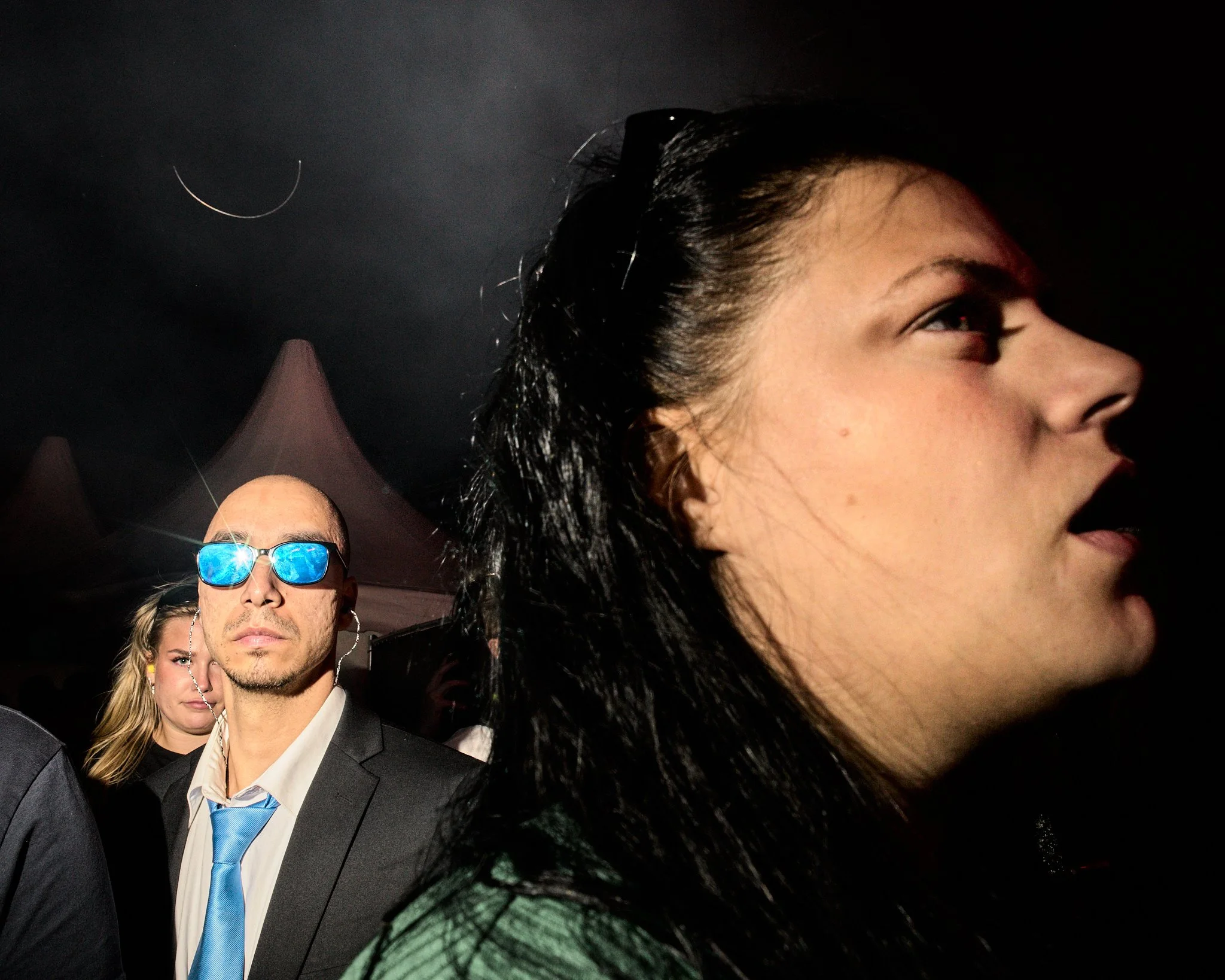 Close-up of a woman with black hair in the foreground, with a man wearing reflective blue sunglasses, a suit, and a blue tie, and a woman with blonde hair in the background at a nighttime event.