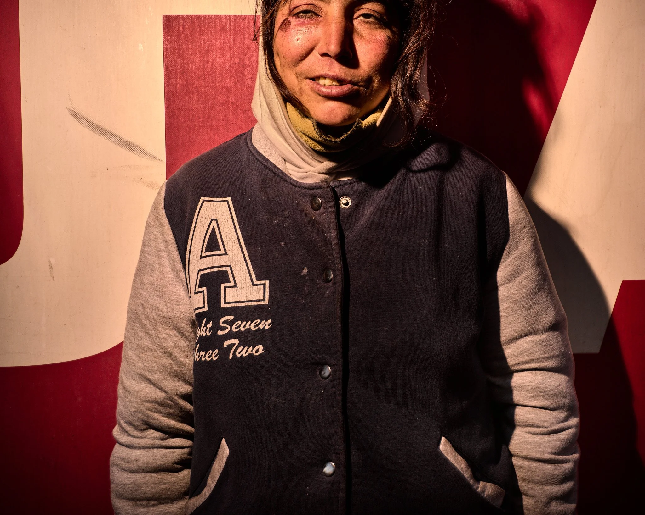 A woman standing in front of a red and white background, wearing a black and gray letterman jacket with an 'A' on it, smiling slightly.