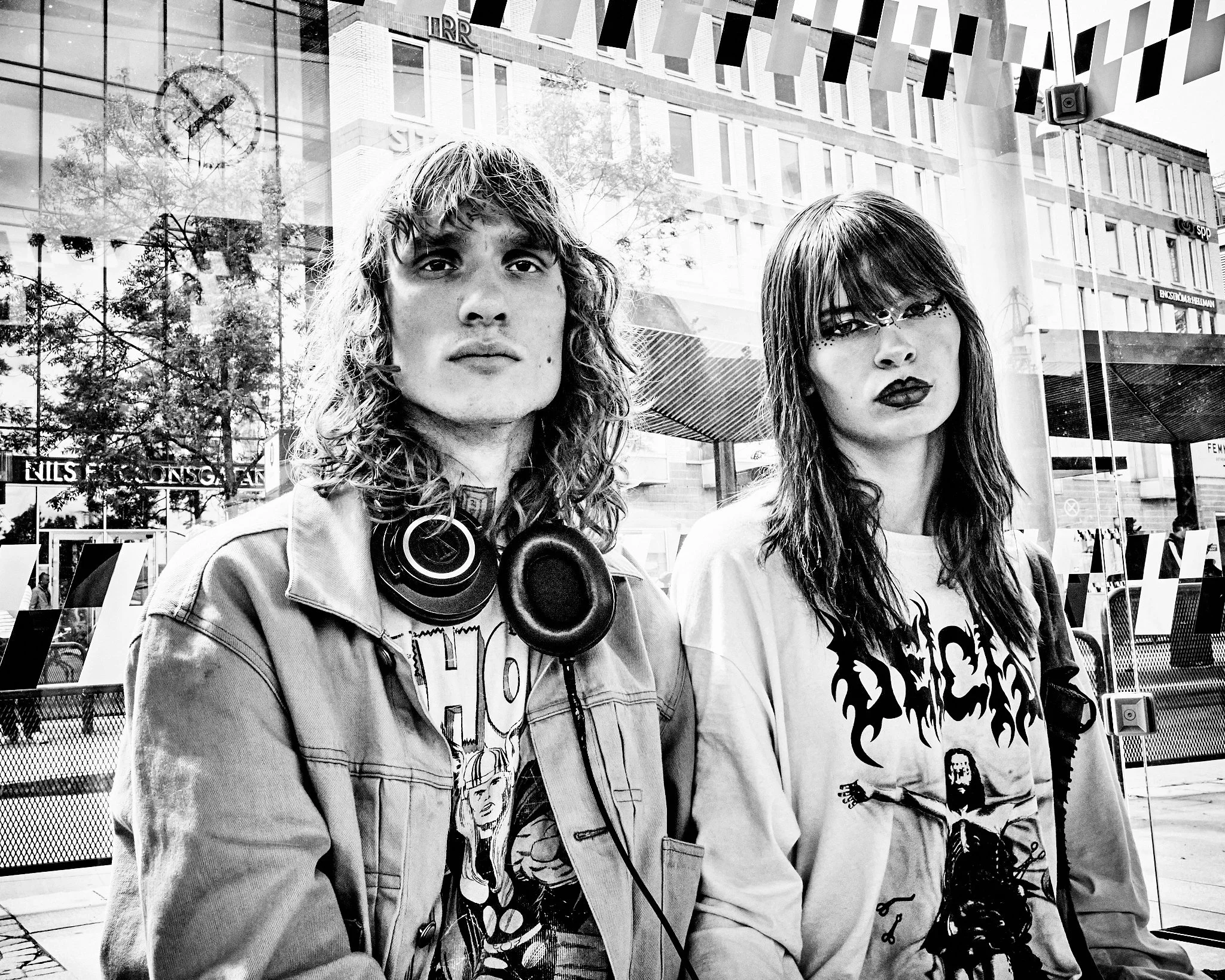 Two young women with long hair and dark makeup pose outside a building, with reflections and city scenery in the glass window behind them.