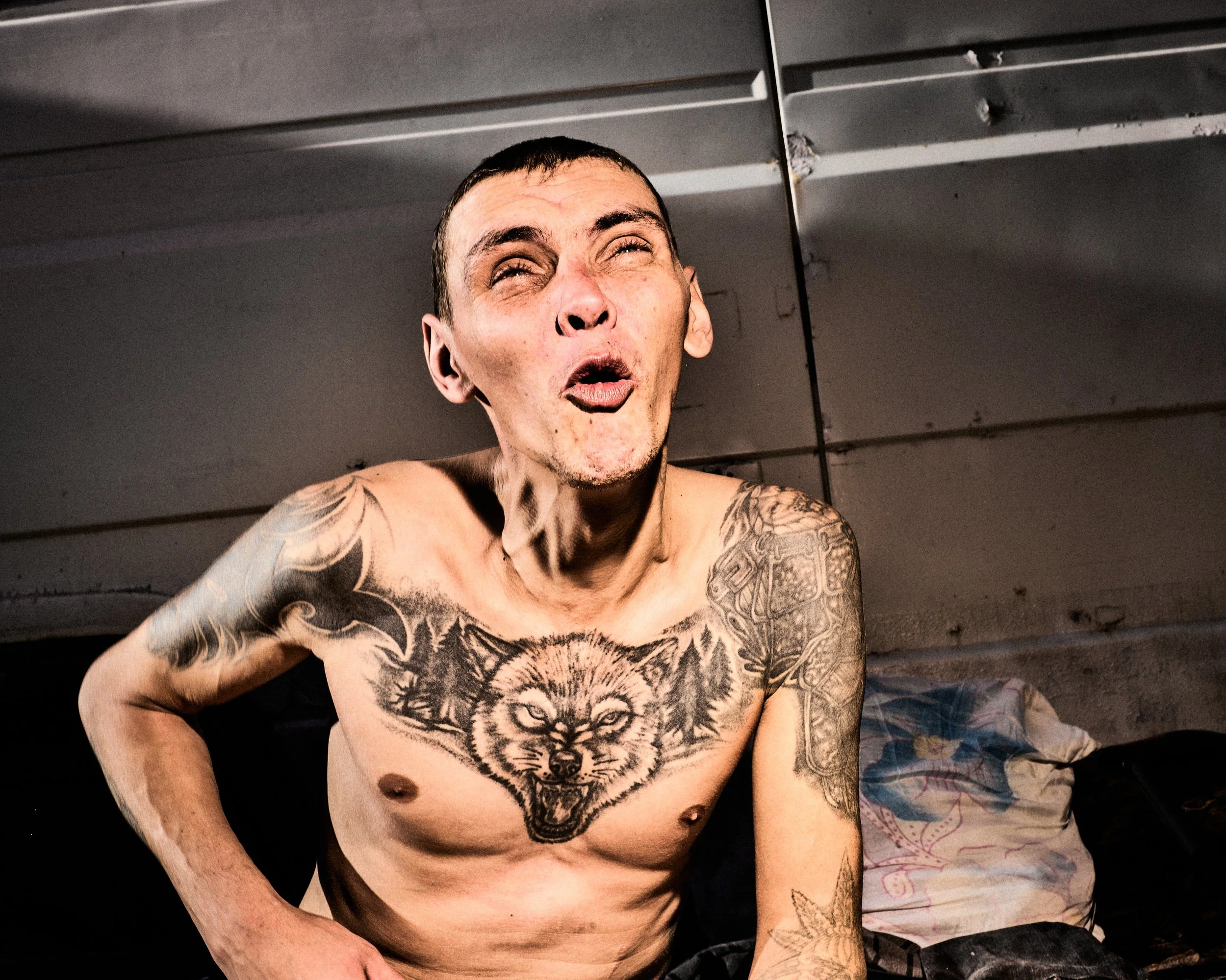 A shirtless man with tattoos, including a large wolf face on his chest, making an expressive face in a room with a metal ceiling and a person partially visible sitting next to him.