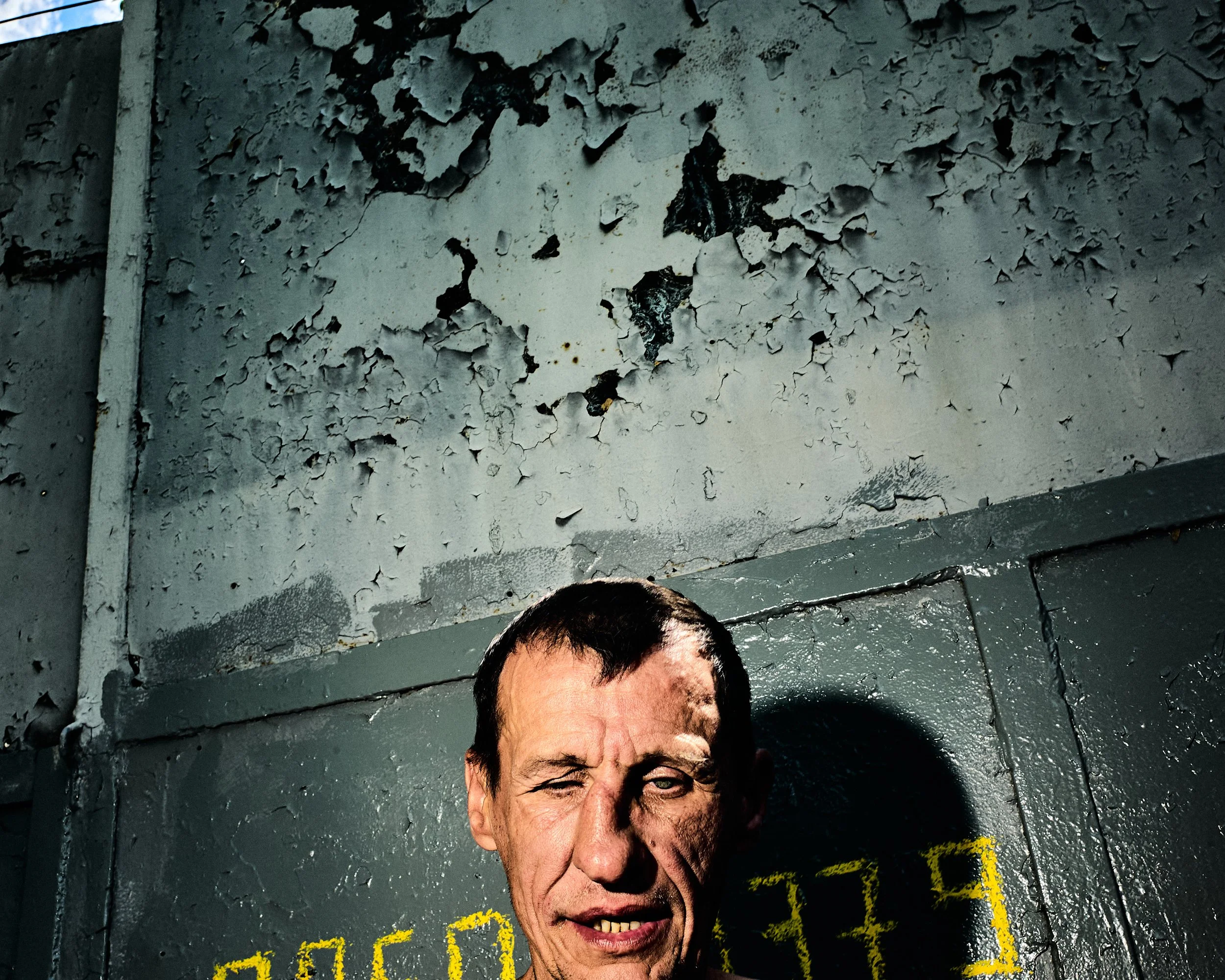 A person with dark hair and a cheerful expression is standing in front of a gray, weathered wall with peeling paint. There is yellow and green text on the wall behind them.