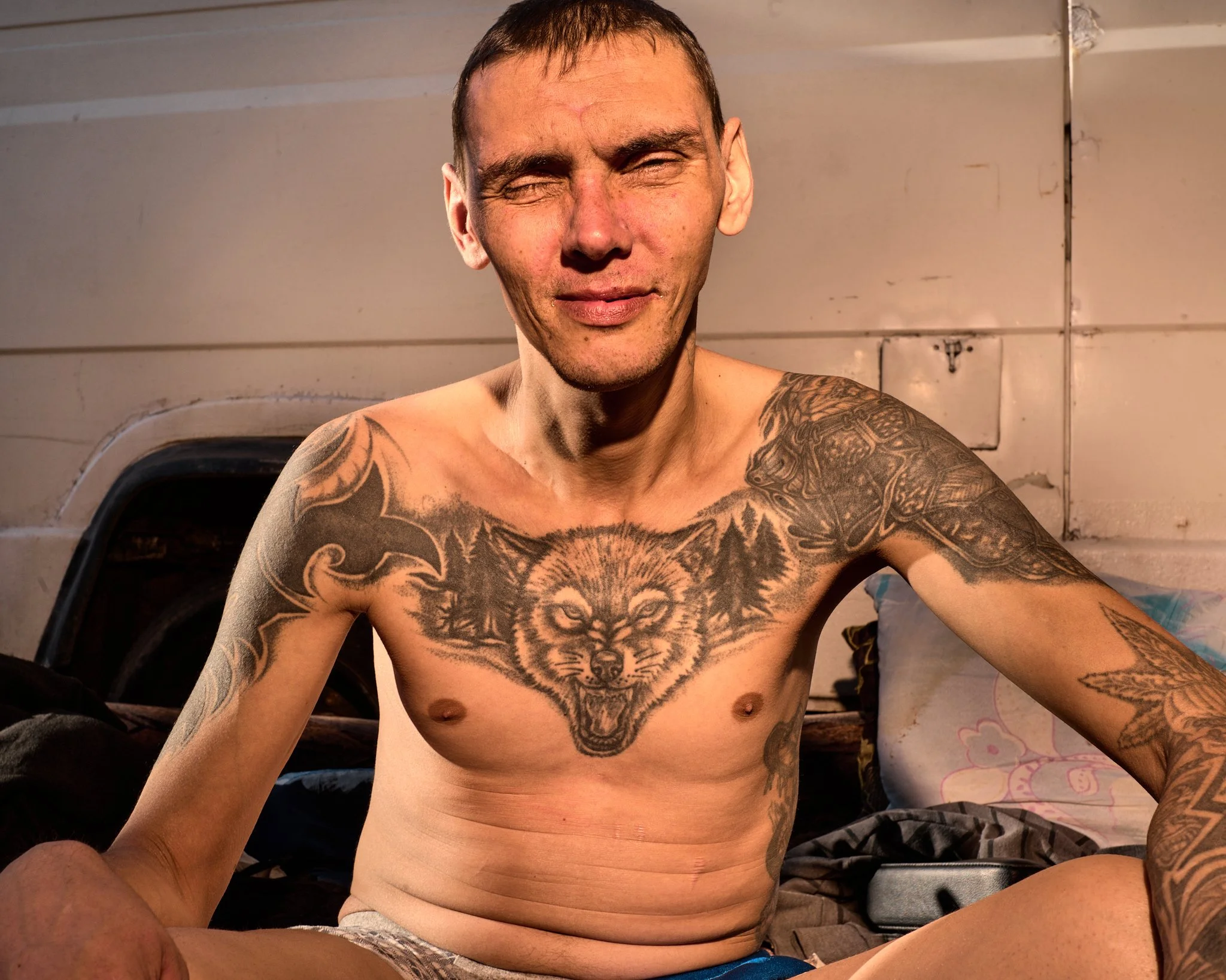 A shirtless man with short brown hair and tattoos sitting in a room with a neutral background.