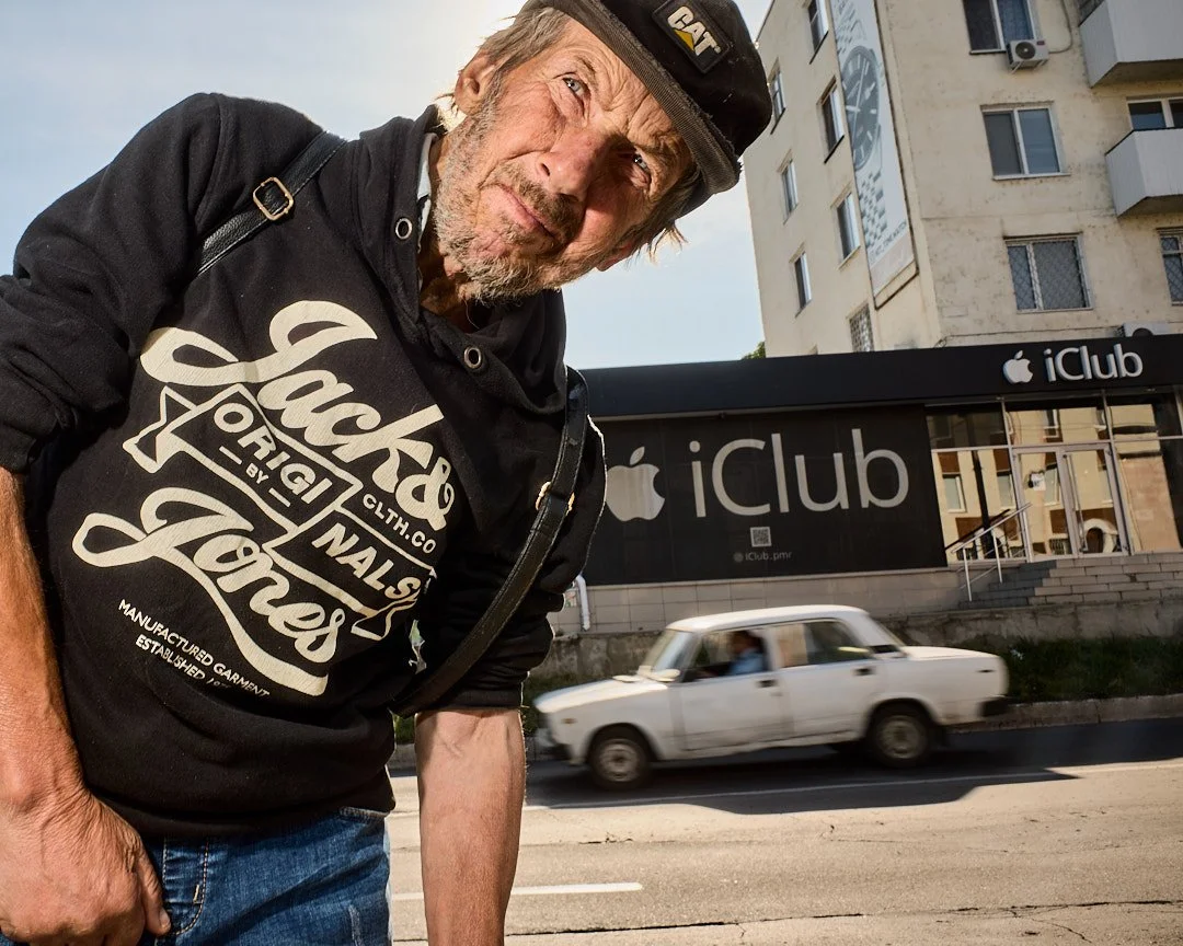 An elderly man with a beard and cap looking at the camera, standing in front of an Apple iClub store with a white older model car passing by on the street.