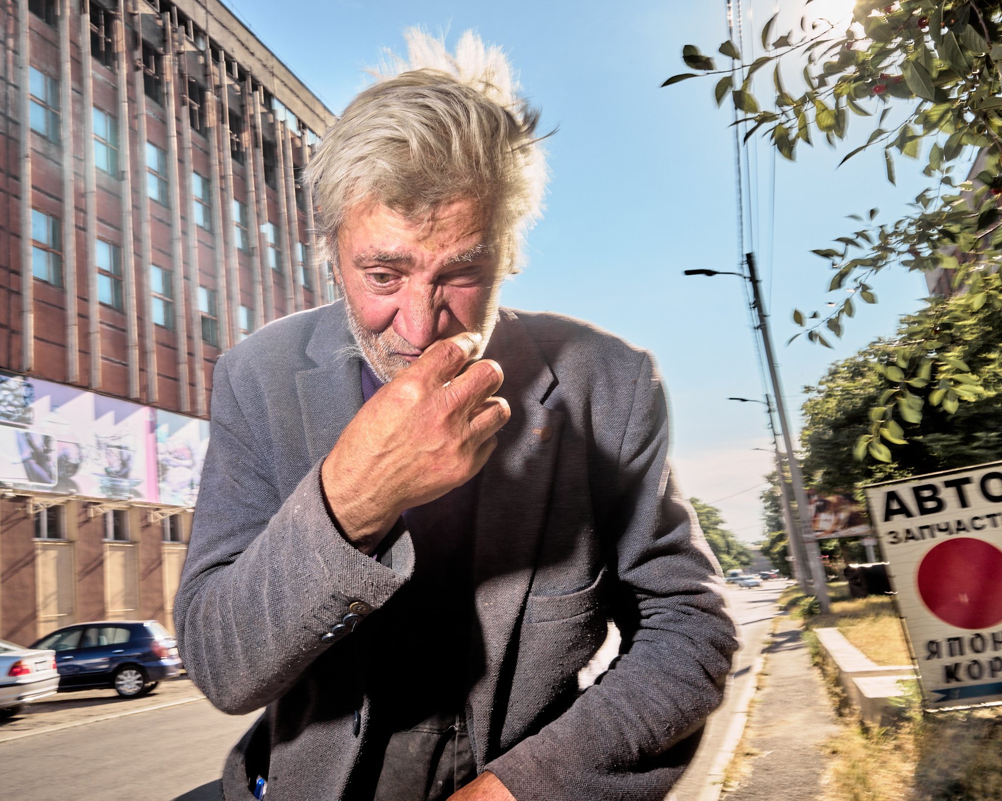 An elderly man with gray hair and beard in a gray suit appears to be sneezing or coughing outdoors on a city street, with trees, telephone poles, and a sign with Russian writing in the background.