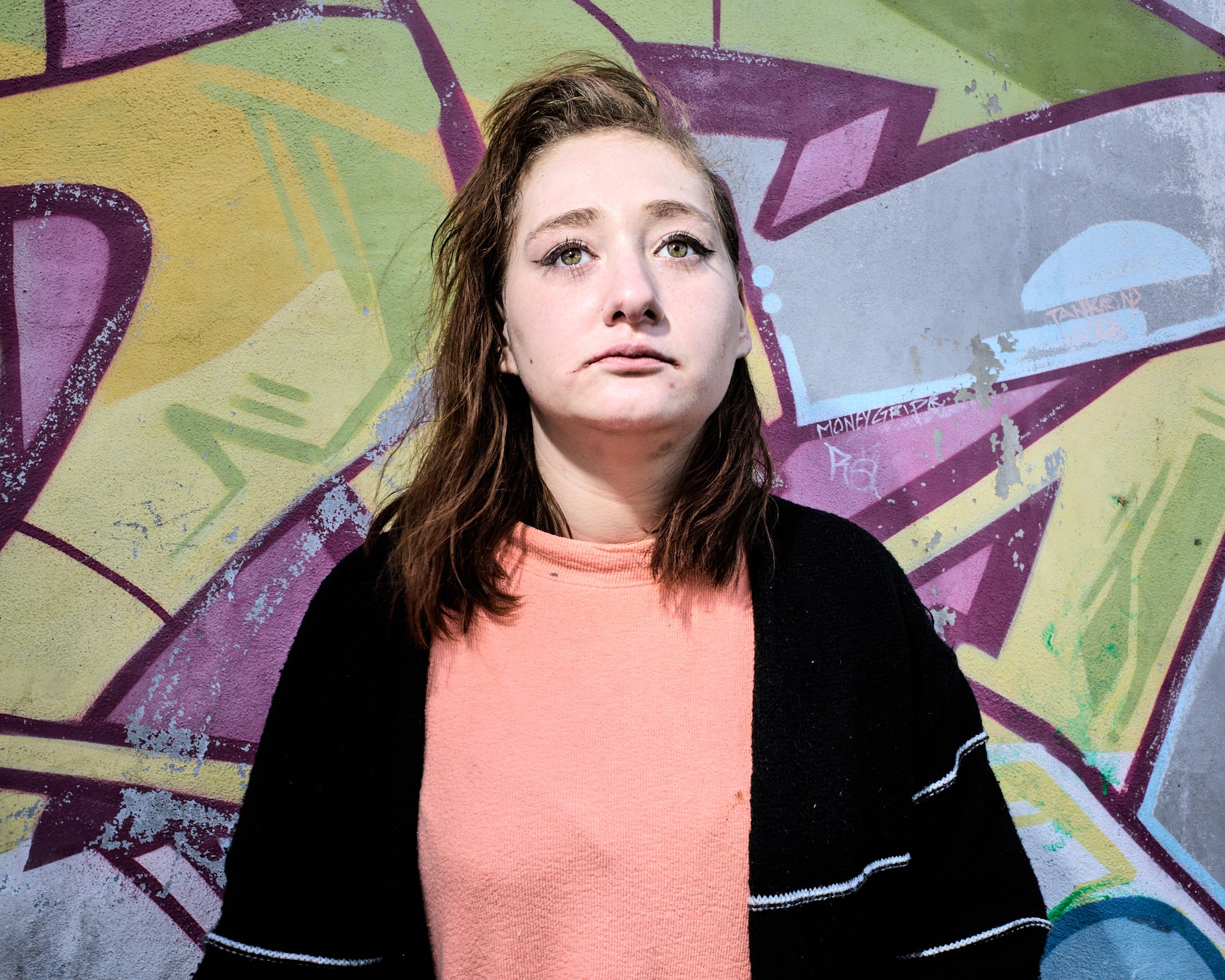 A young woman with shoulder-length brown hair, wearing a pink shirt and black sweater with white stripes, standing in front of a colorful graffiti wall.