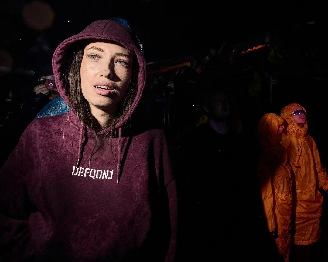 Woman wearing a dark red hoodie with 'DIE.FQON.1' printed on it, standing in a dark environment with other people, some wearing orange rain gear.