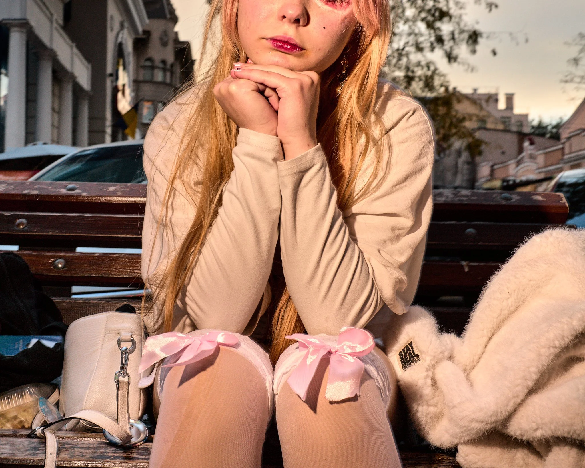 A young woman with long red hair seated on a park bench, wearing a beige jacket and pink knee-high stockings with bows, resting her chin on her clasped hands. She is outdoors during the evening, with buildings and trees in the background.