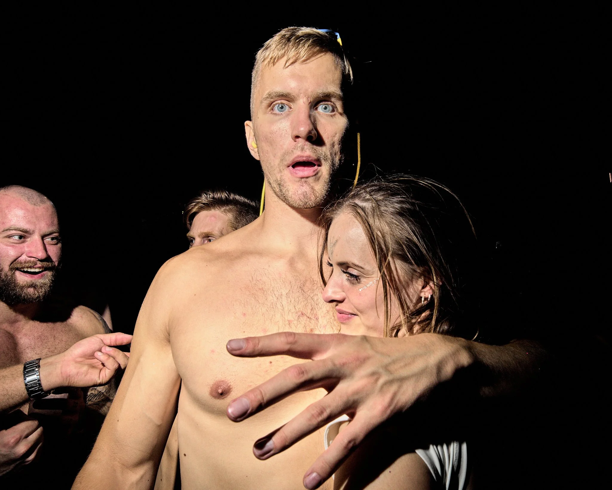 Shirtless man and woman with other shirtless men in the background, on a dark background, celebrating or engaging in an intense moment.
