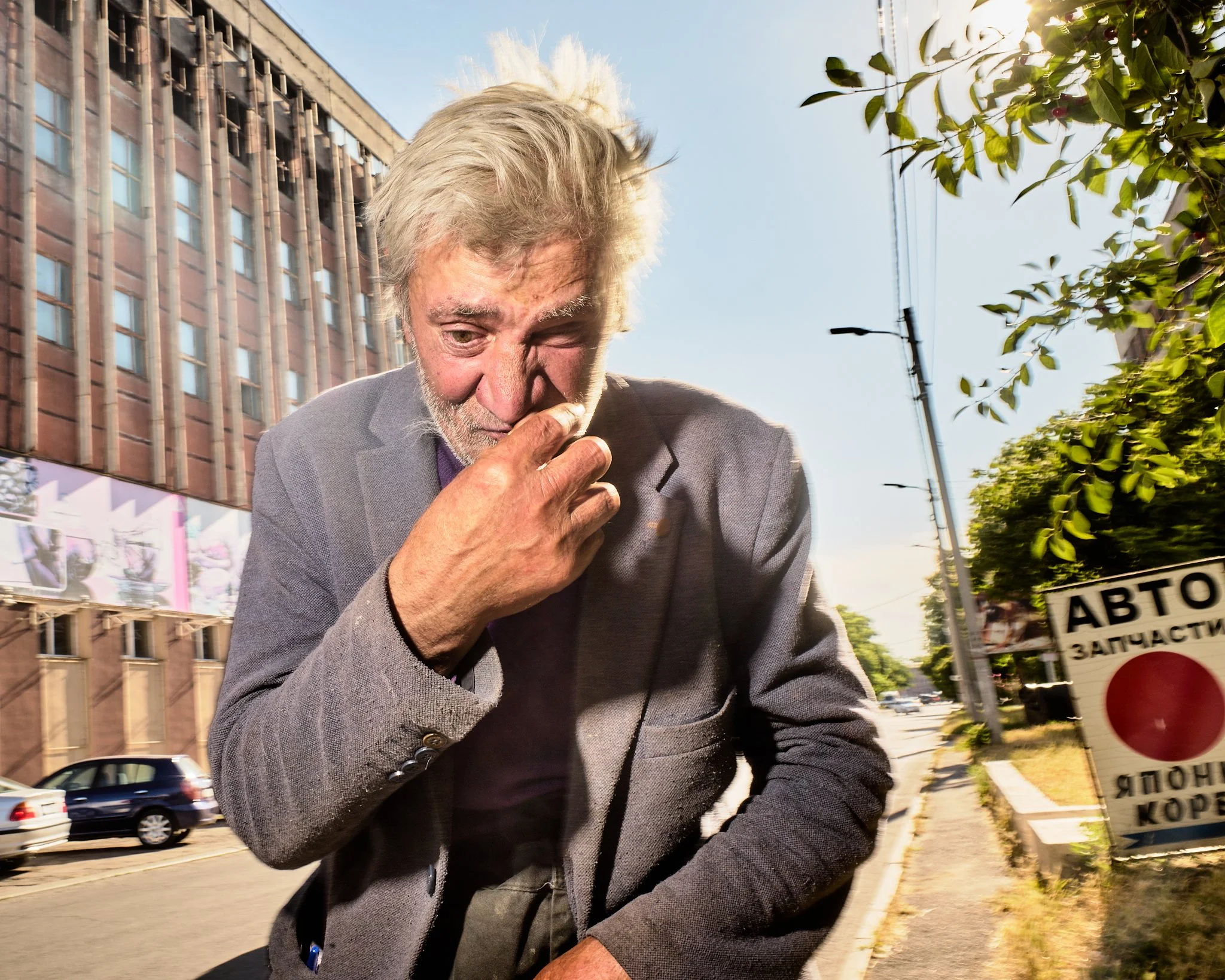 An older man with gray hair and a beard, dressed in a gray blazer, is standing on a city street, smelling or inspecting his fingers, with a worried or distressed expression. The background shows cars, a building, and a sign with Cyrillic text.