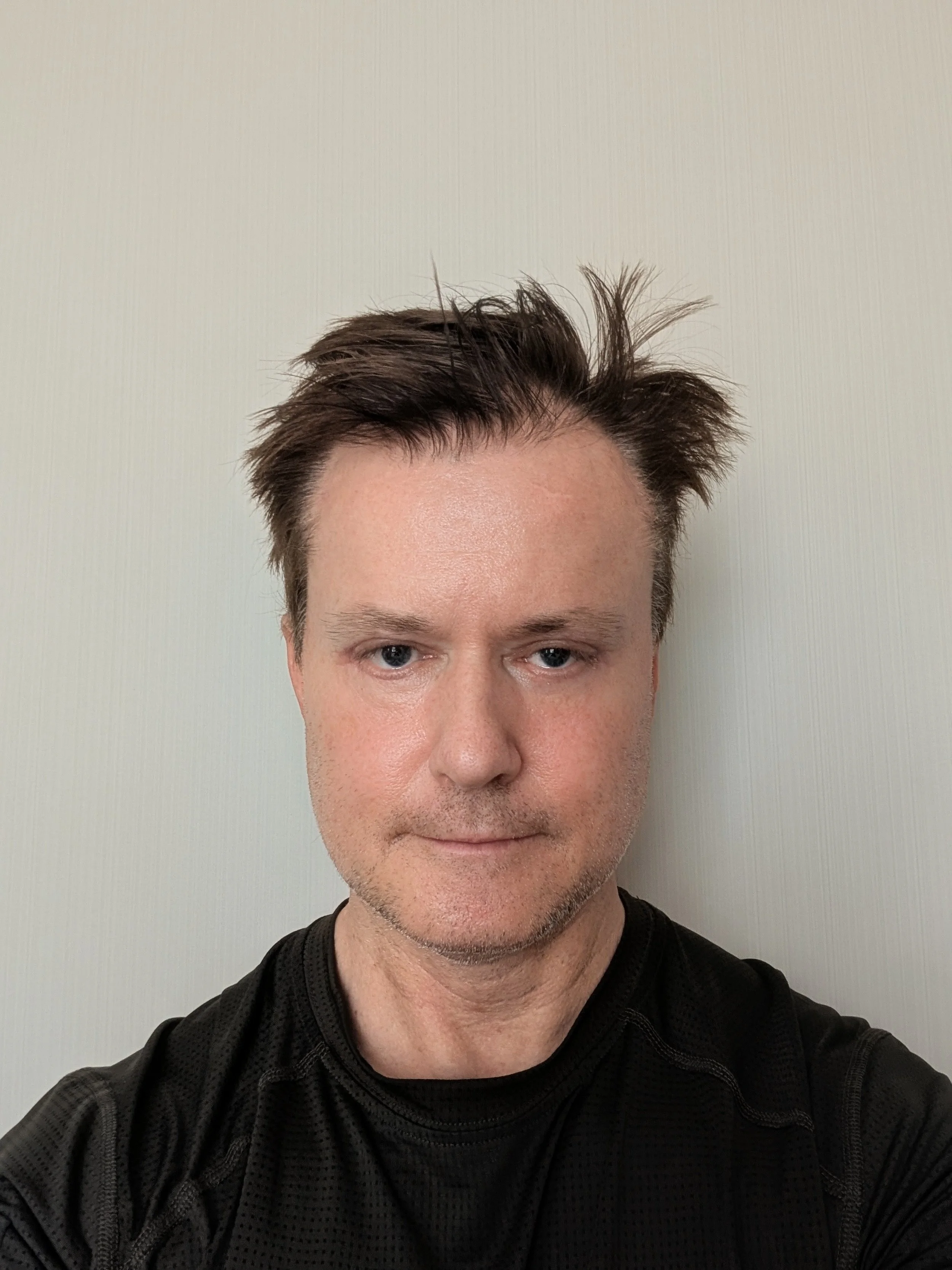 A man with short dark hair and blue eyes taking a selfie against a light-colored wall, wearing a black athletic shirt.