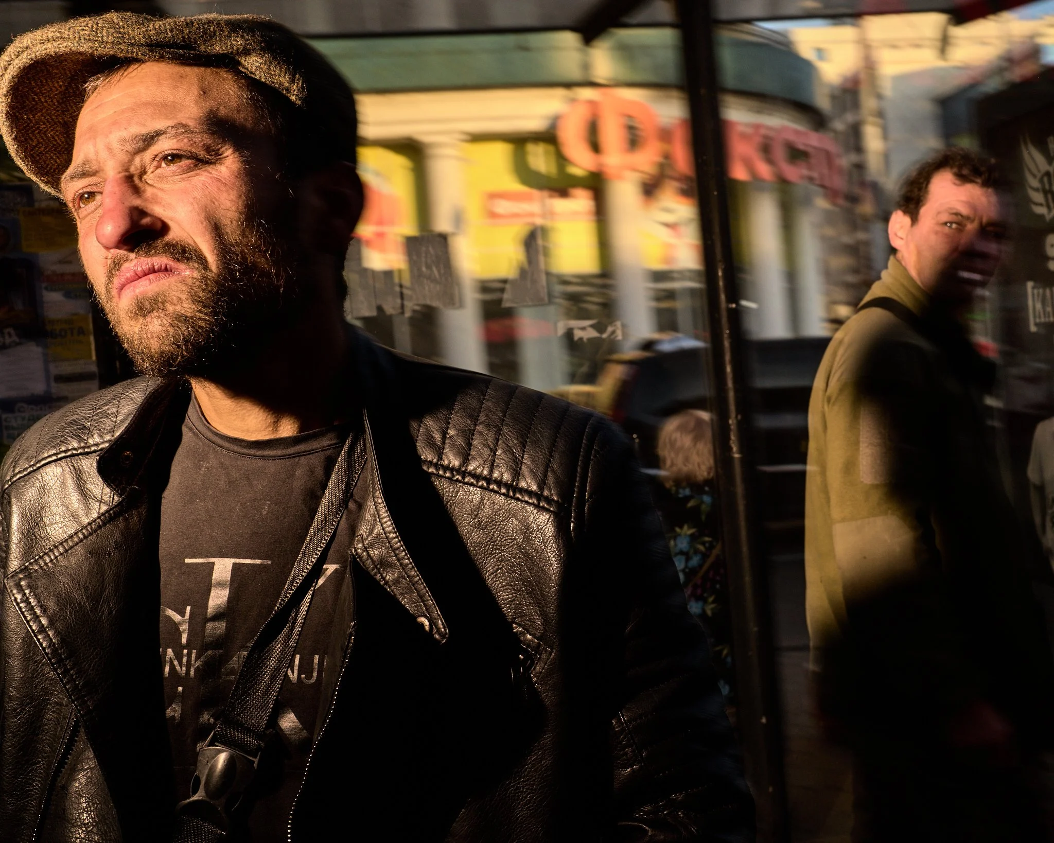 A man with a beard and wearing a leather jacket and a hat, standing outside with sunlight casting shadow on his face, with reflections of city buildings and another man behind a glass window.