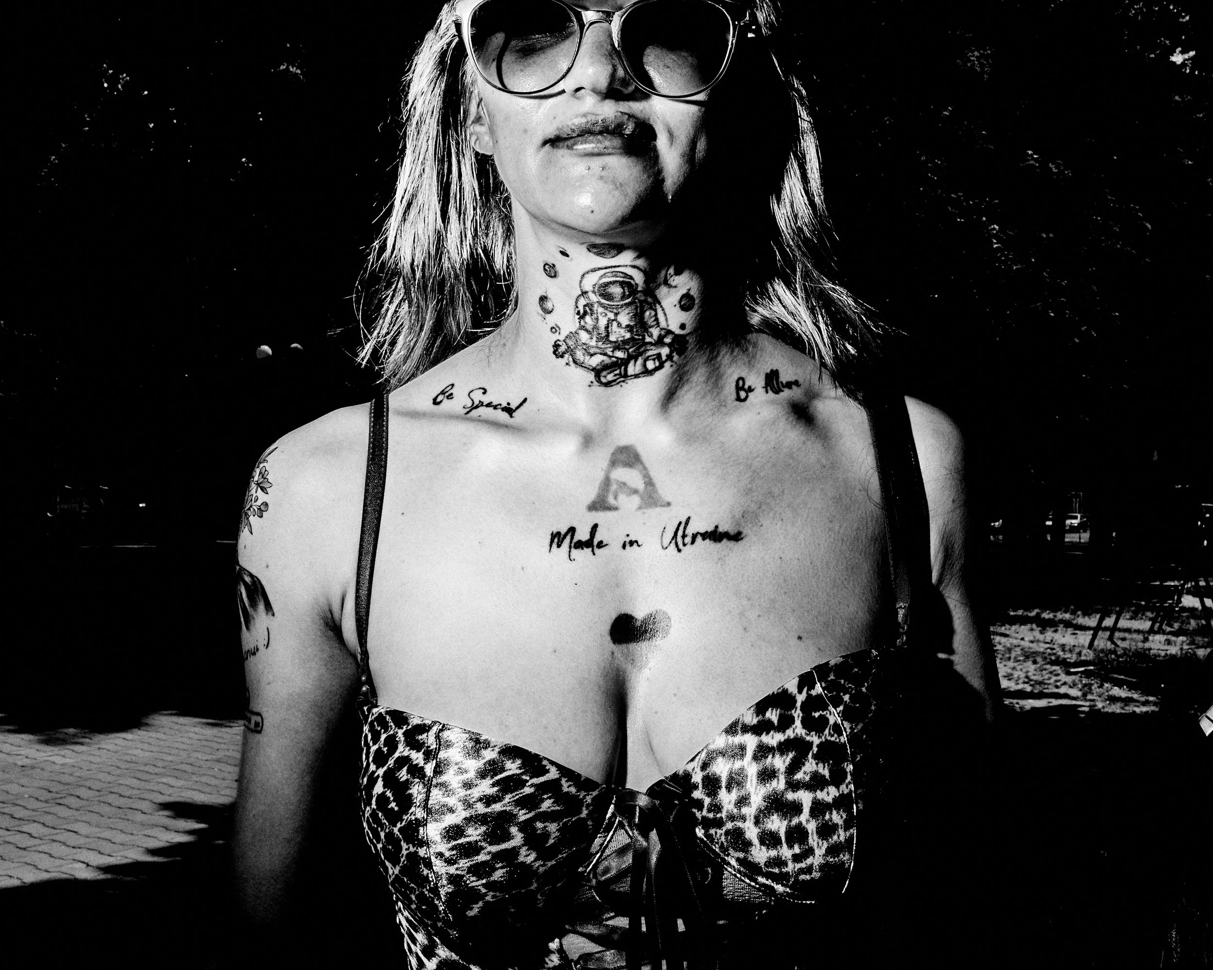 A woman with sunglasses and long hair, tattoos on her neck and chest wearing a leopard print outfit, standing outdoors in sunlight.