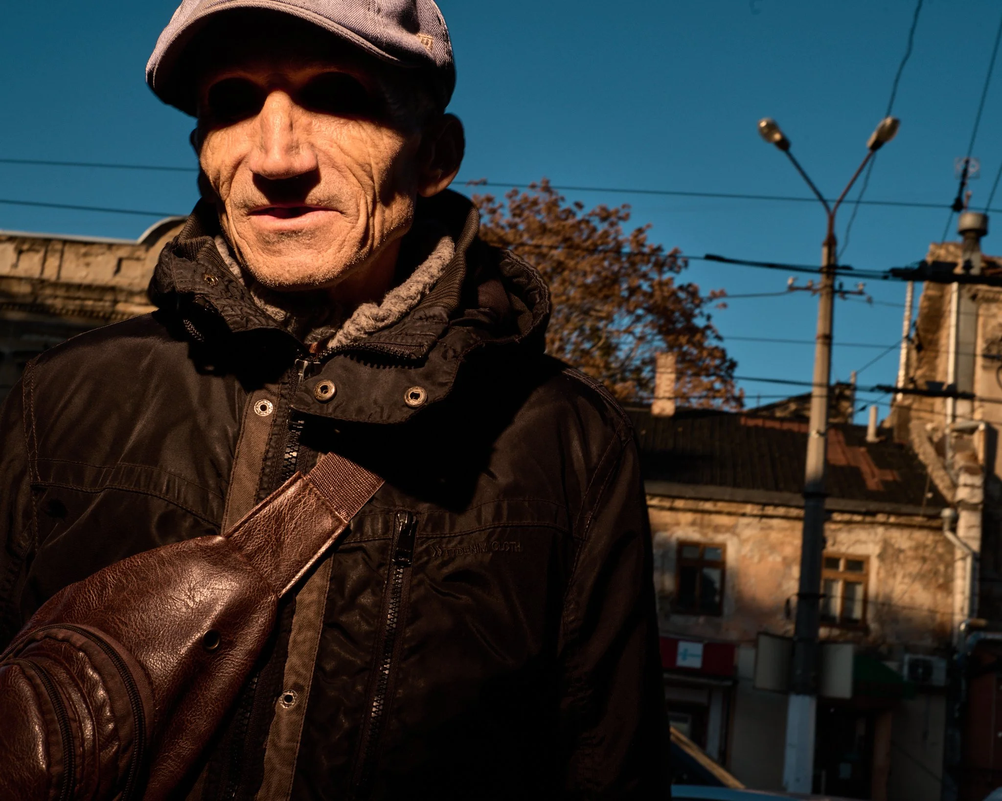 An elderly man in a black jacket and a brown leather bag across his shoulder, wearing a hat and a gray hoodie layered underneath, standing outdoors in front of buildings, power lines, and a blue sky during daytime.