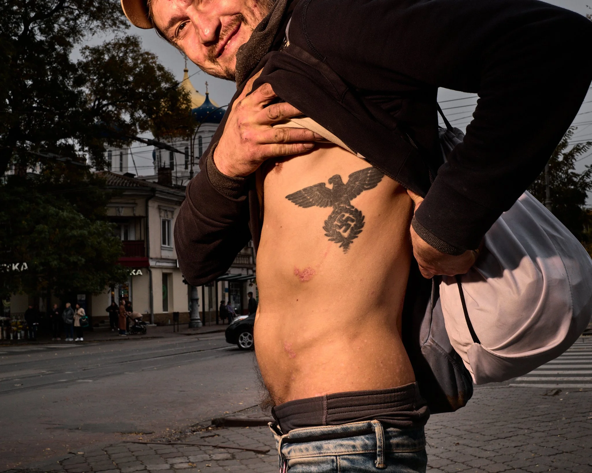 A man with a smile is lifting his shirt to reveal a tattoo of a Nazi eagle with swastika on his chest, while standing on a city street.