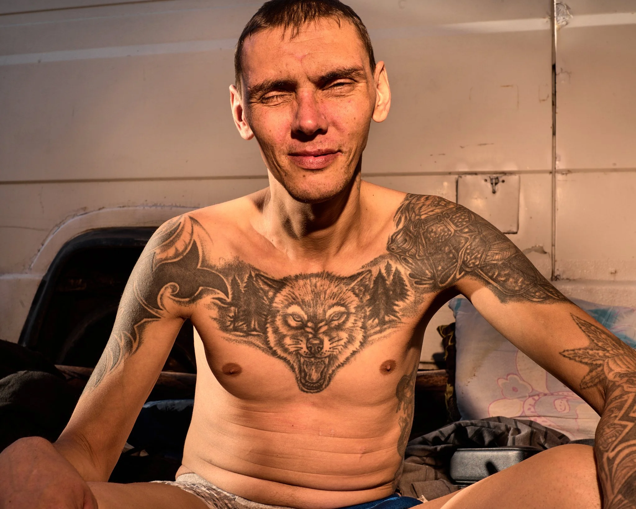 A shirtless man with tattoos, sitting and smiling, in front of a background with a wall and some blankets or clothing.