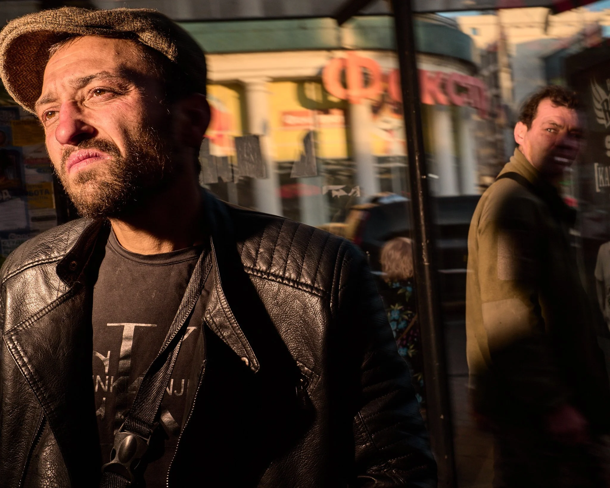 Two men standing outside in warm sunlight with a storefront reflected behind them. The man on the left is wearing a leather jacket and a hat, while the man on the right is in a jacket, both looking in different directions.
