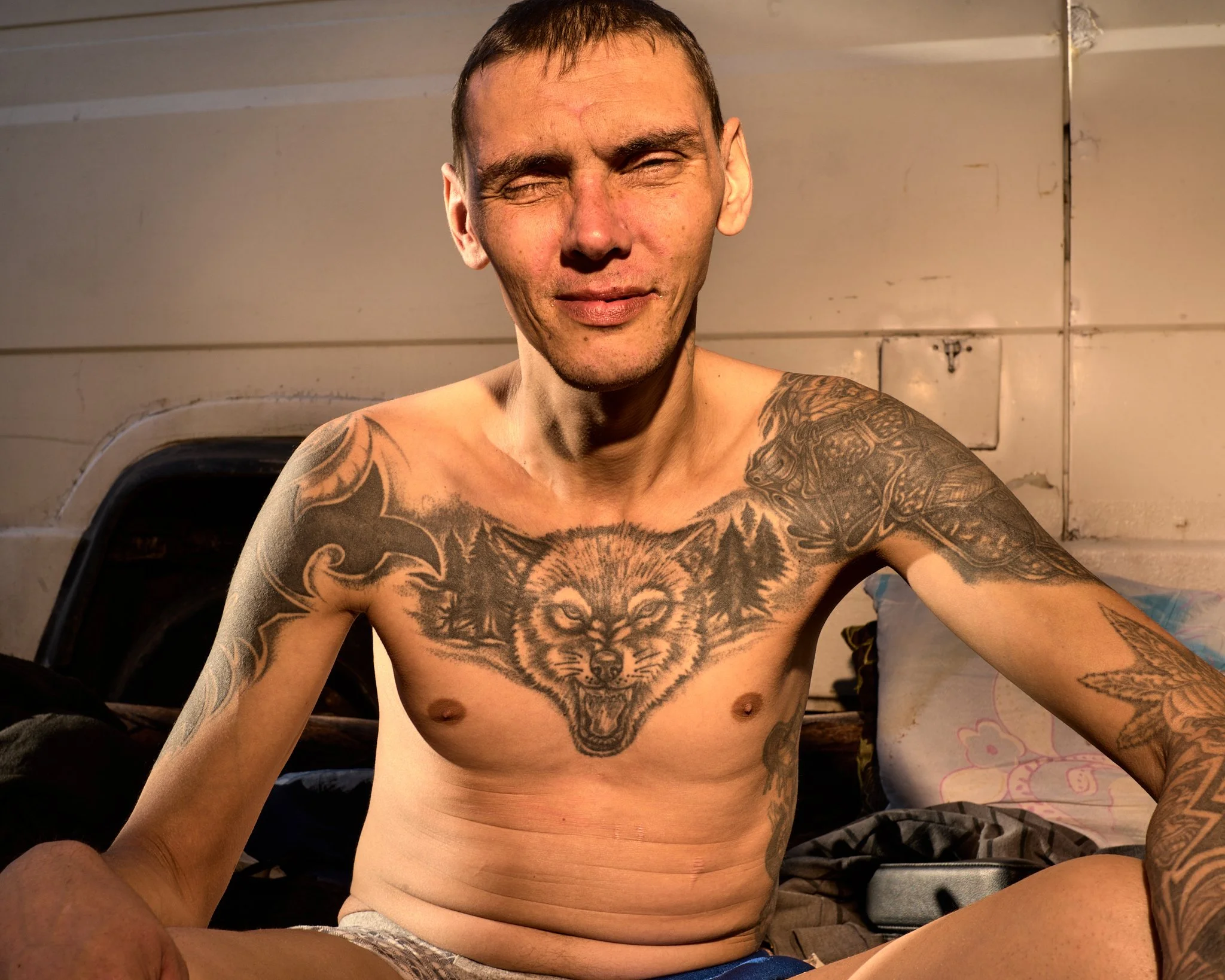 A shirtless man with tattoos sitting on a bed, smiling, with a wooden wall background.