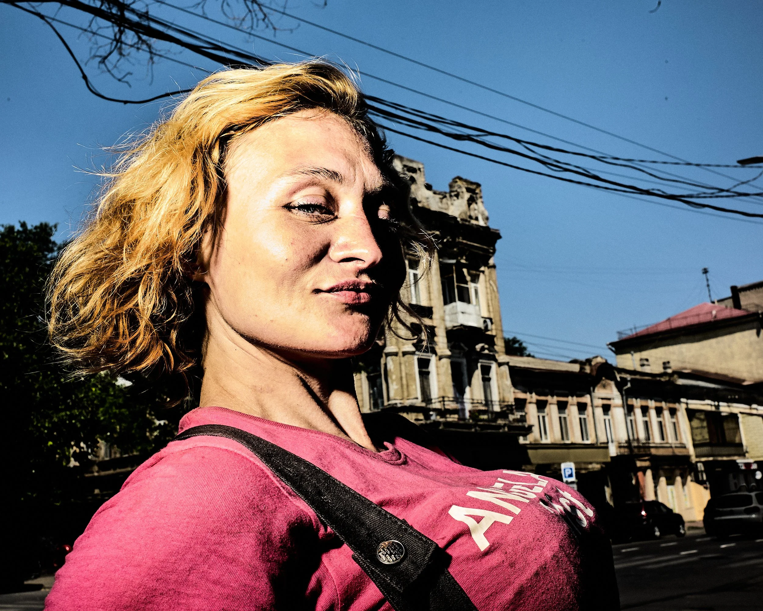 A woman with blonde curly hair wearing a pink shirt, carrying a black strap bag, standing outdoors on a sunny day with blue sky. There are old buildings and power lines in the background.