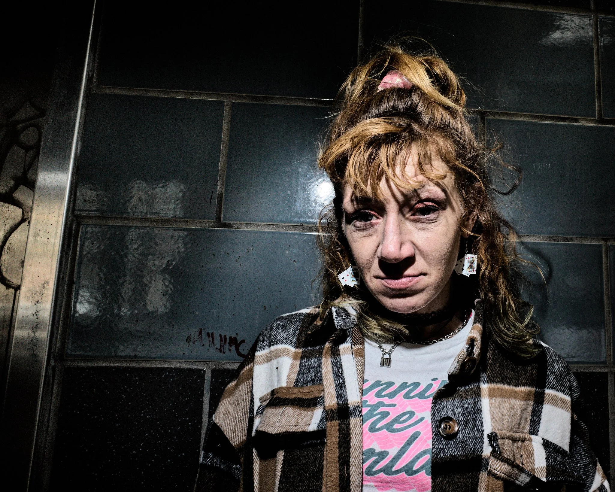 A woman with curly, reddish hair styled in a high messy bun, wearing earrings, a plaid jacket, and a graphic T-shirt, standing against a dark tiled wall.