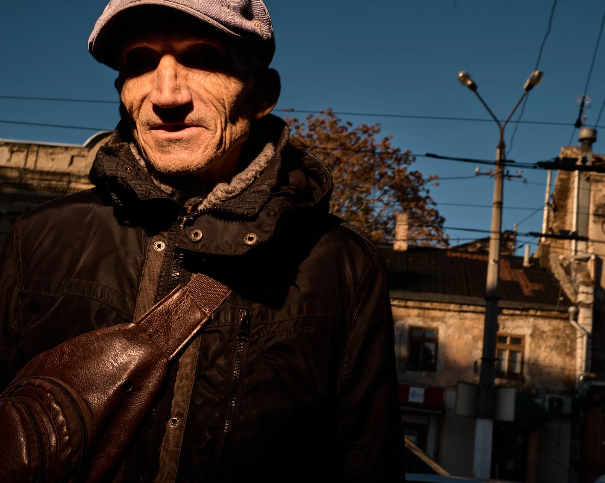 Close-up of an elderly man wearing a gray hat and dark jacket, with sunlight illuminating his face, against an urban background with buildings, power lines, and a tree.
