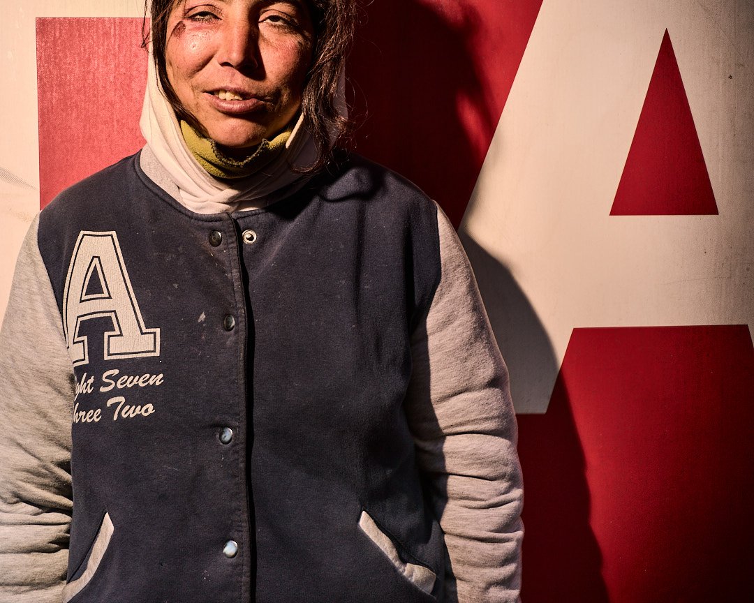 A woman with dark hair wearing a black and gray varsity jacket standing in front of a red and white background.