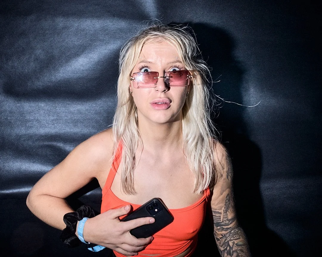 A young woman with blonde hair wearing pink-tinted sunglasses and a bright orange top, holding a phone in her hand, with a surprised or confused expression on her face.