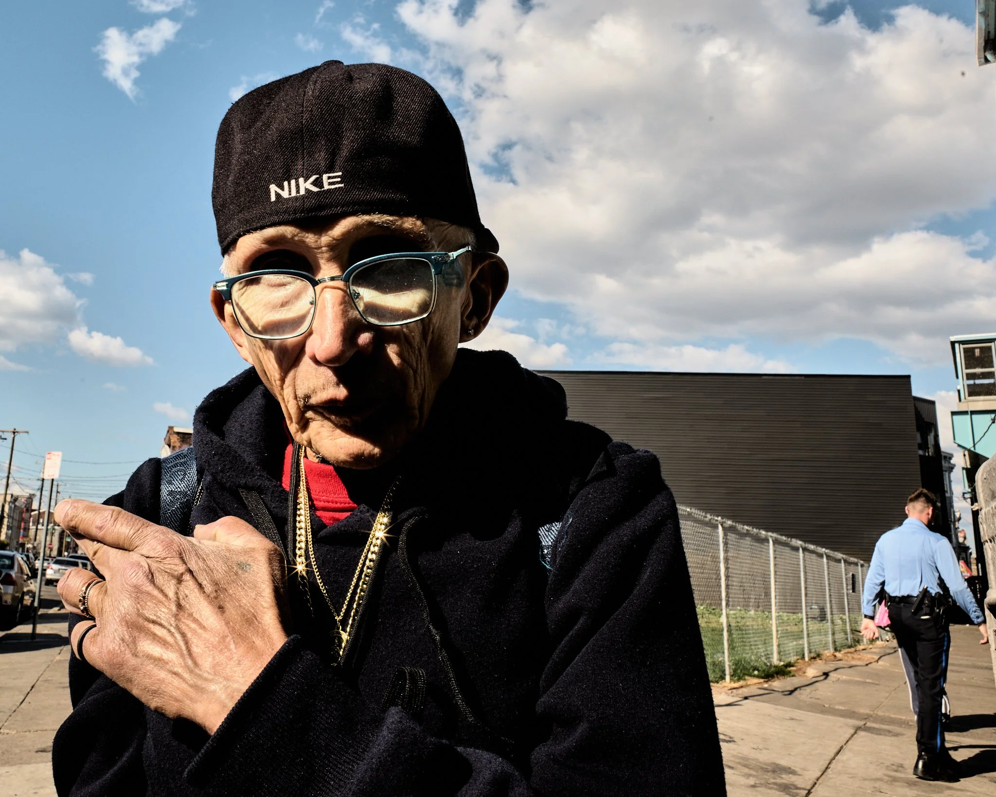 Close-up of an elderly man with glasses, wearing a black Nike cap, gold chains, and a black hoodie, standing outdoors on a sunny day with a partly cloudy sky, with a man in a police uniform walking away in the background.
