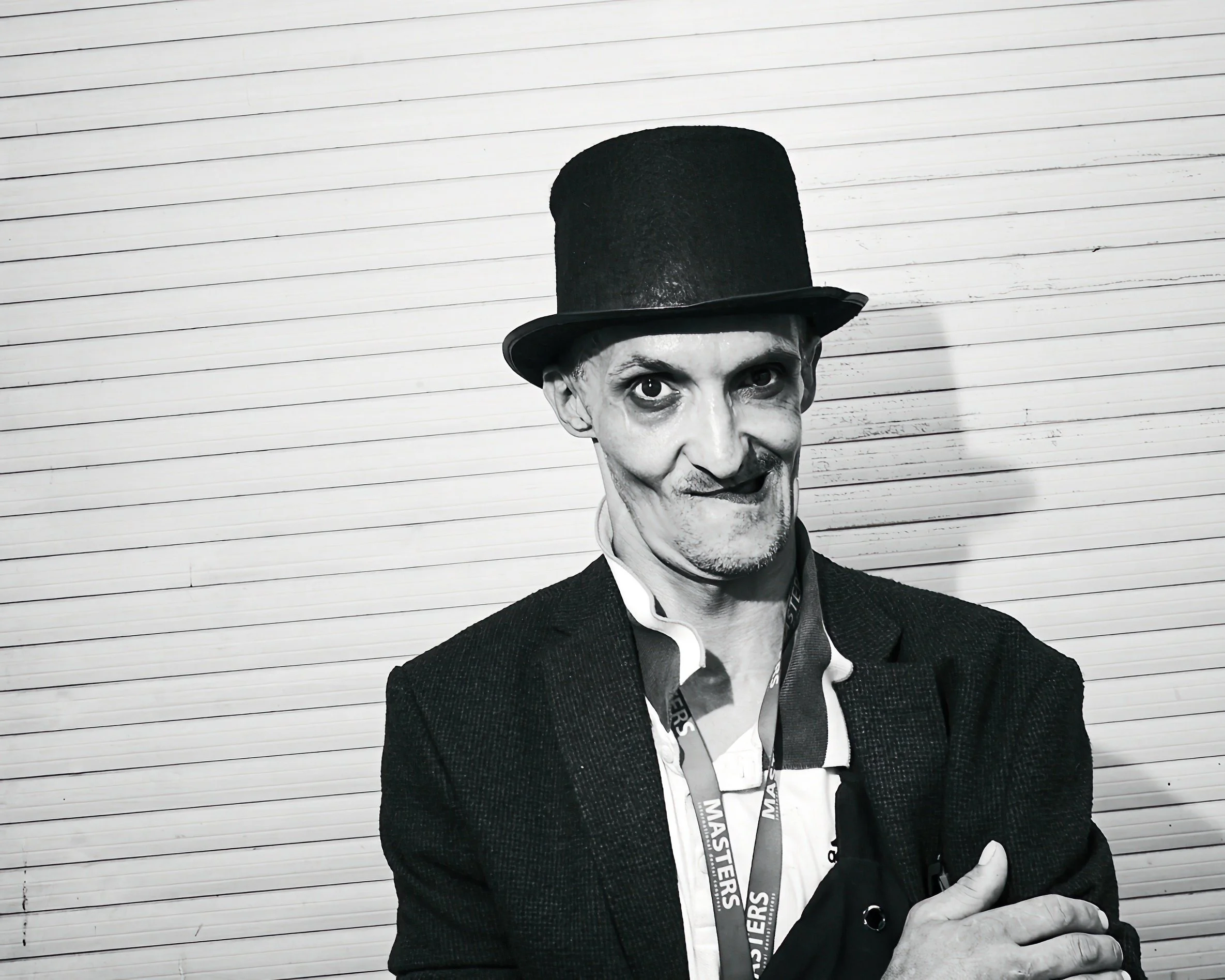 A person dressed as a mime with a painted face, wearing a black top hat, a suit, and a lanyard, standing in front of a white wooden wall.